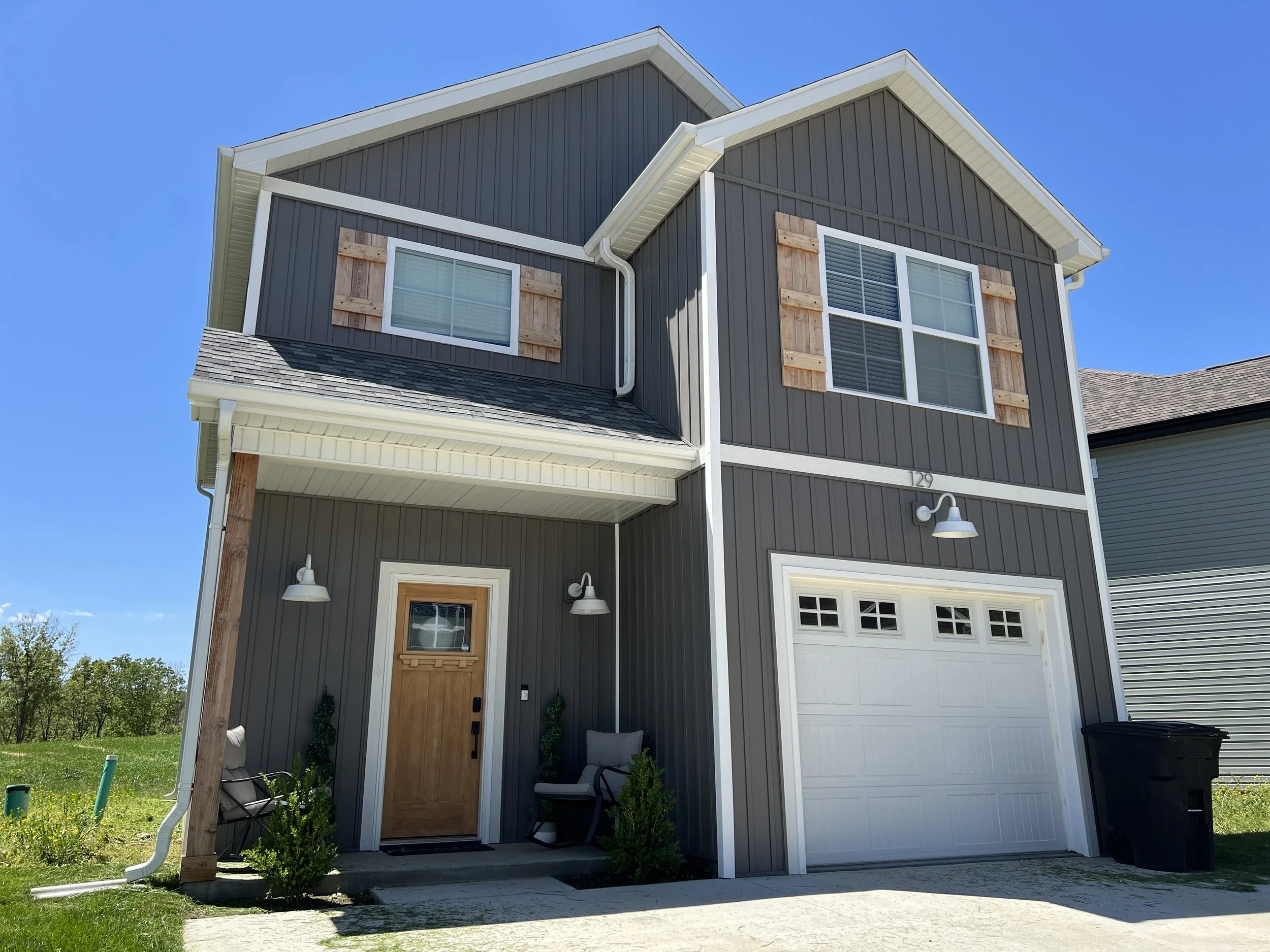 Two-story modern house with dark gray siding, white trim, and a wooden front door. Features include shingle roof, exposed wooden shutters, a white garage door, outdoor light fixtures, and some greenery near the entrance. Clear blue sky in the backgro