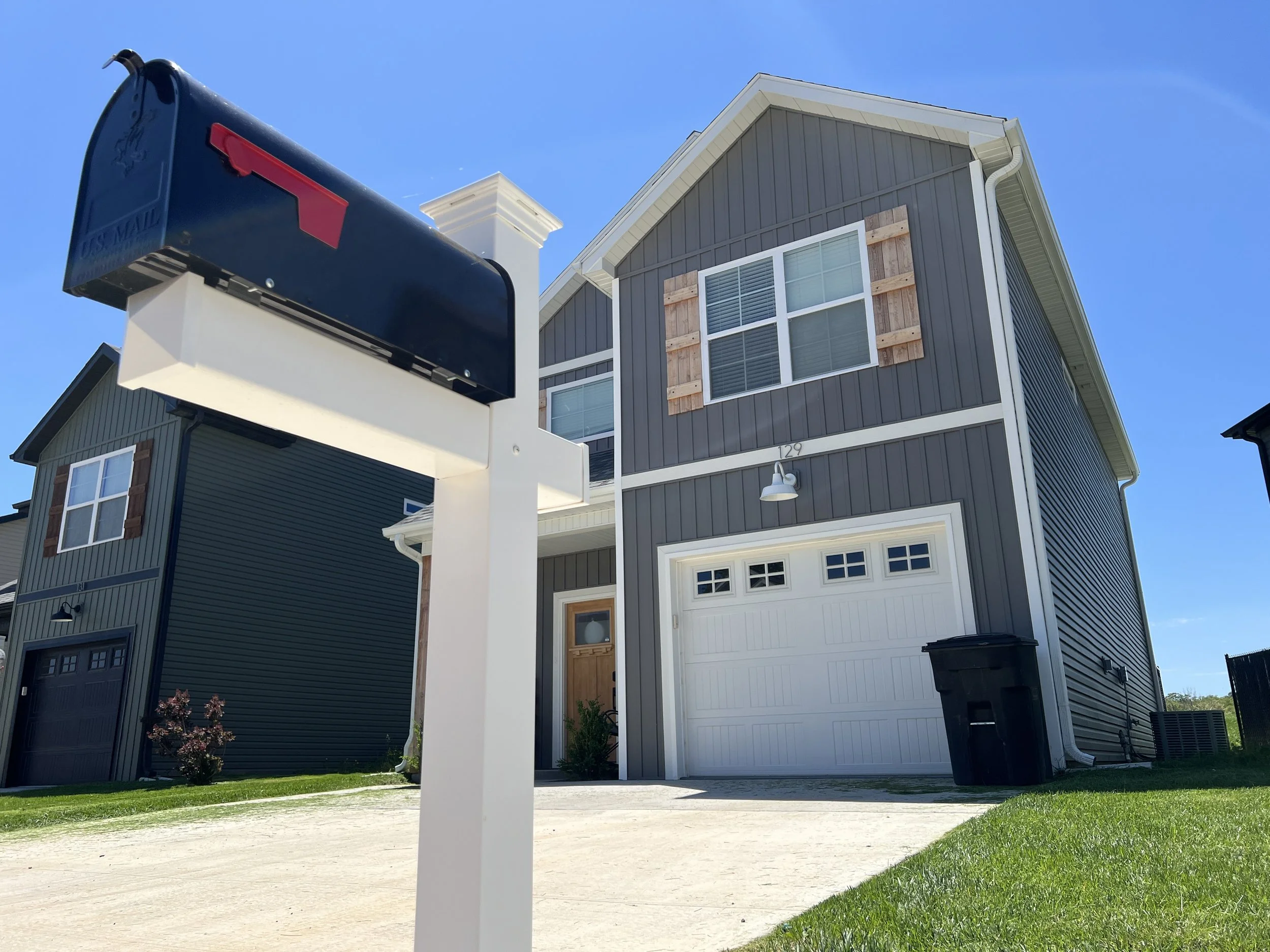 Front view of a modern two-story house with gray siding, white garage door, and a small front yard with green grass. A mailbox is in the foreground.