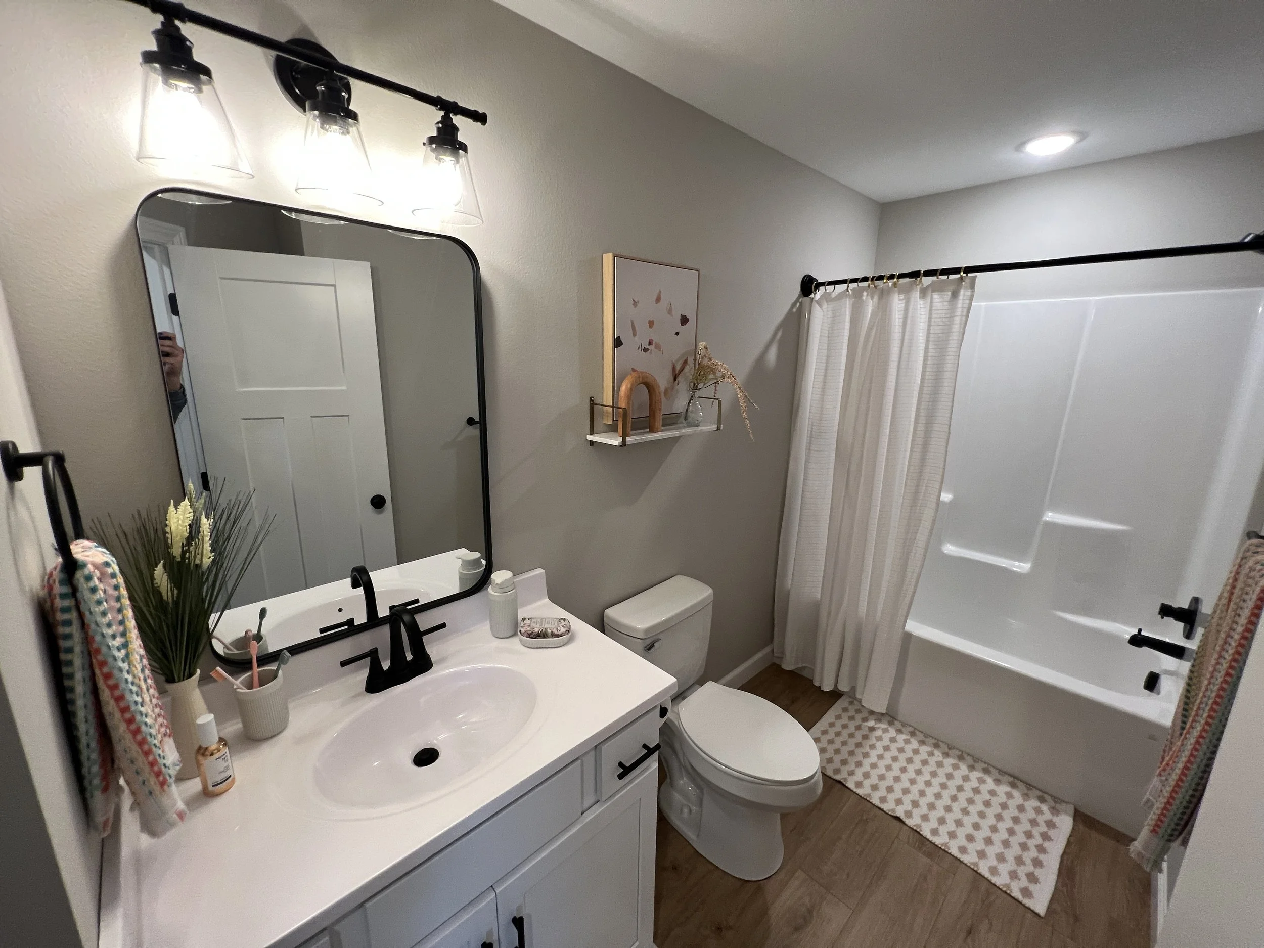 A bathroom with a white vanity, black fixtures, a large mirror, a toilet, a bathtub with white shower curtain, wooden flooring, and decorative items including a framed art, small shelf, plants, and towels.