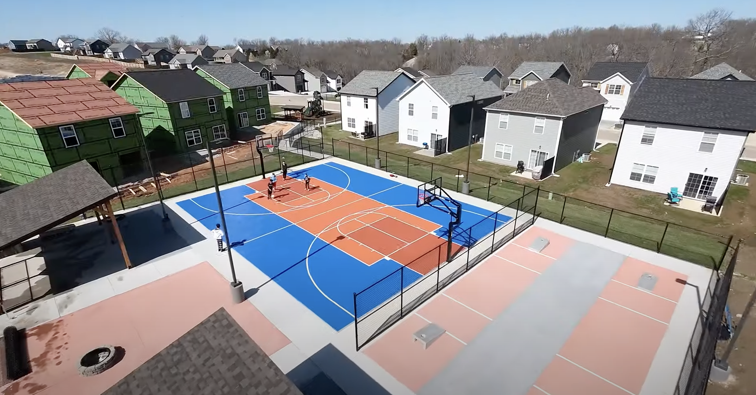 Aerial view of a residential neighborhood with new construction houses and a sports complex featuring a blue and orange basketball court and a smaller multi-sport court.