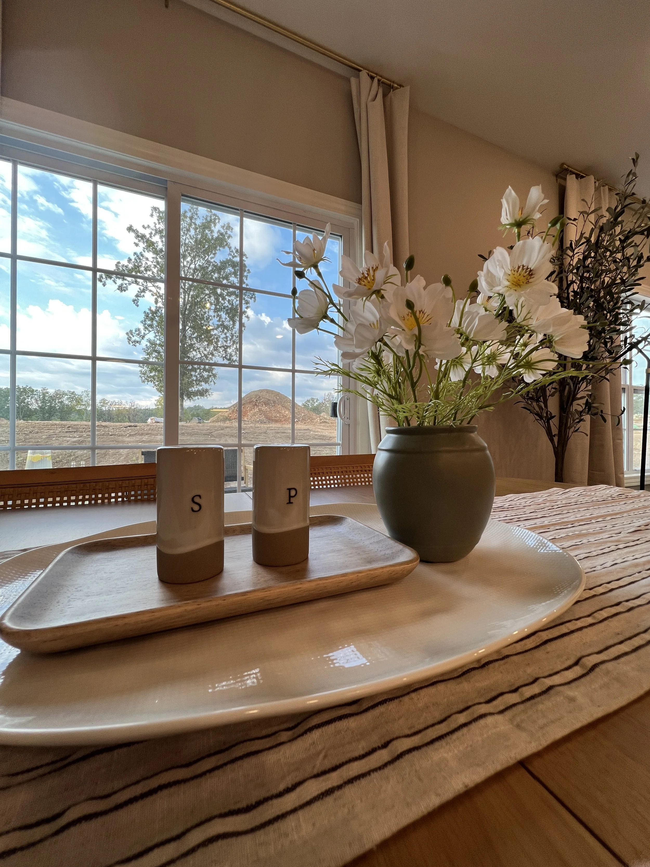 A dining table with a large beige and white striped table runner, a white platter, a wooden tray with two salt and pepper shakers labeled 'S' and 'P', and a vase with white flowers, with a window showing a blue sky, trees, and a dirt mound outside.
