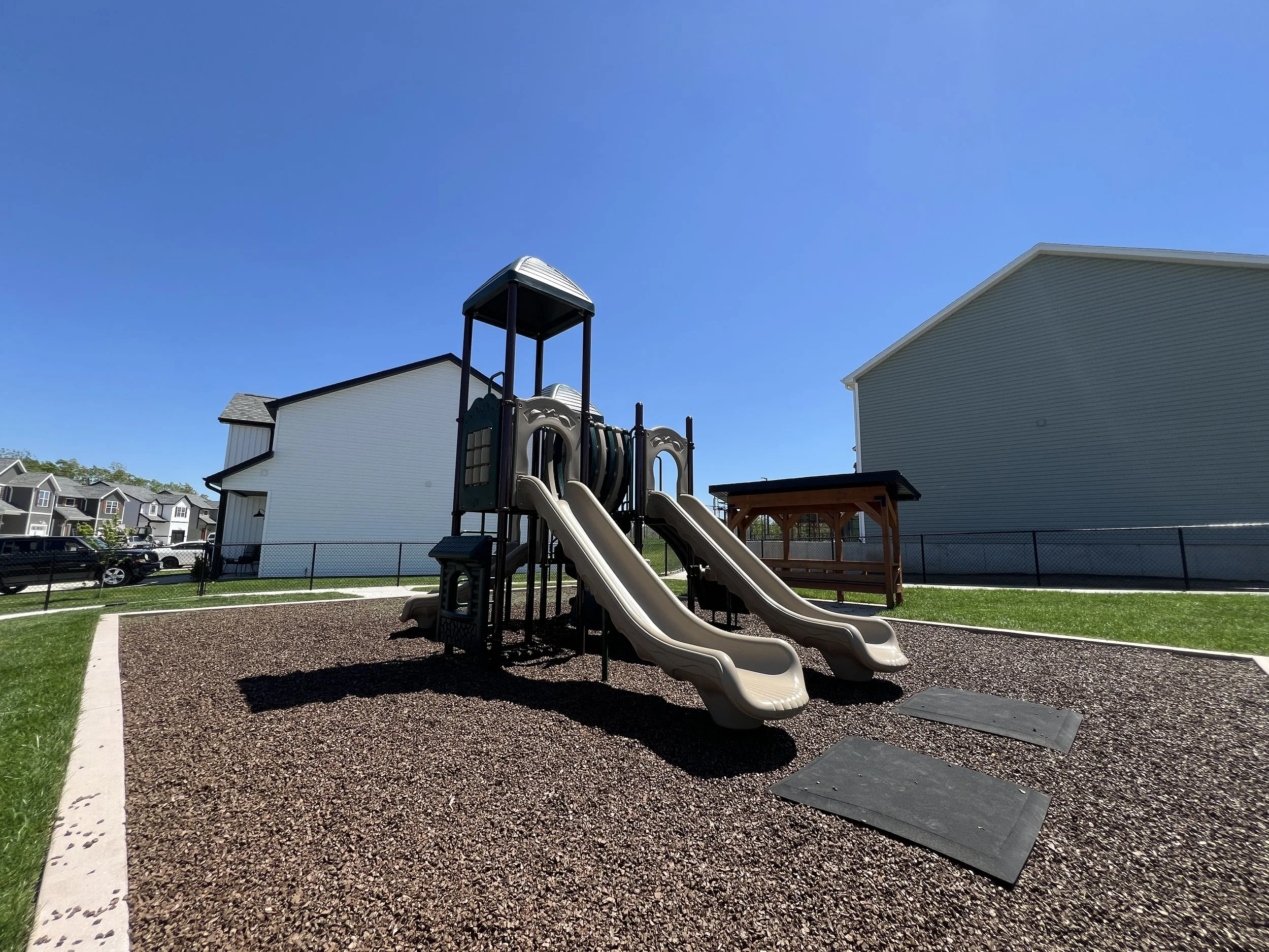 Playground with two beige slides, black support structure, and rubber mats on gravel surface, surrounded by houses and a clear blue sky.