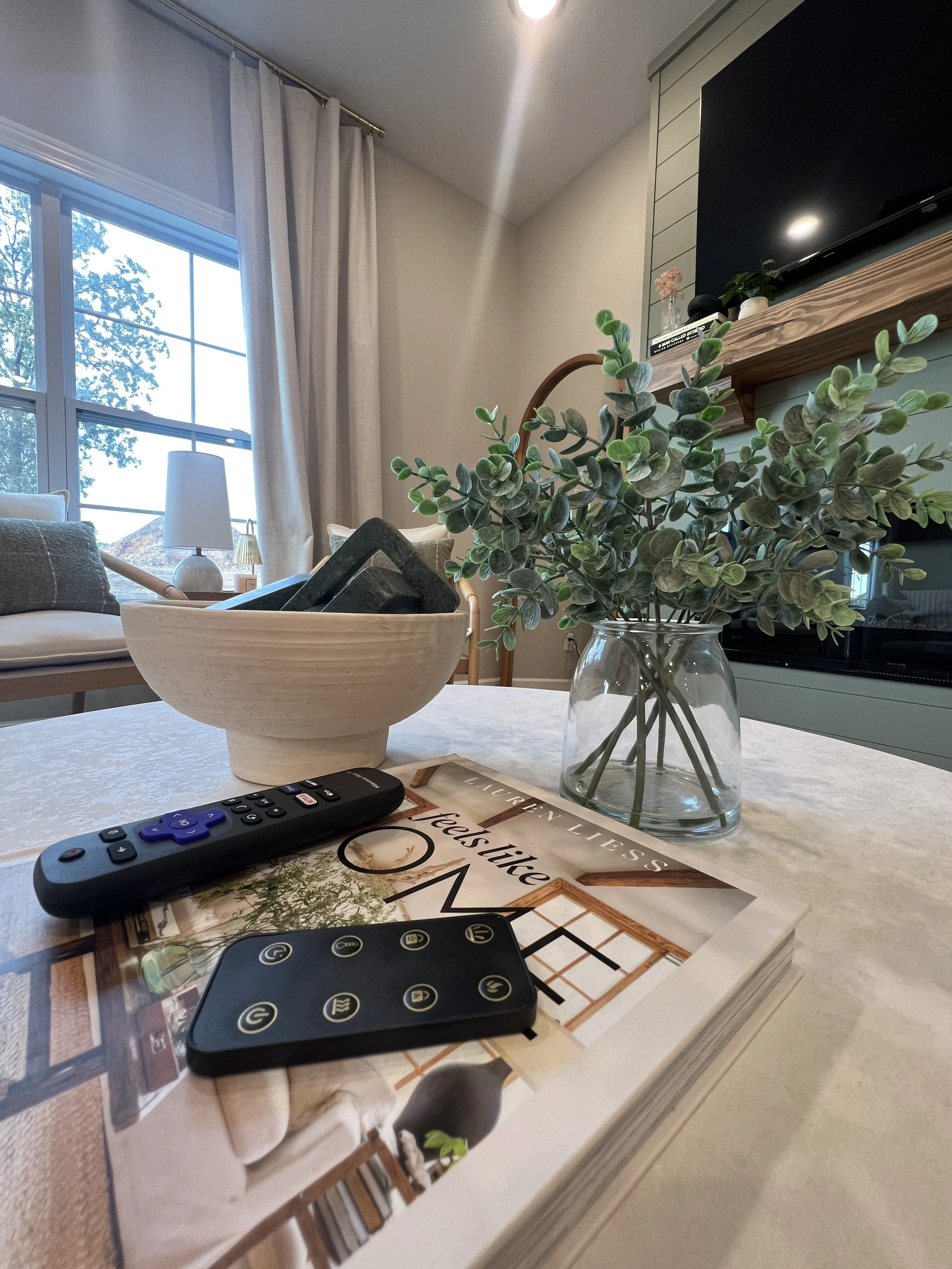 Close-up of a living room table with a remote control, a magazine, a key fob, a glass vase with lush green leaves, a large bowl with black objects, a window with white curtains, and some furniture and decor in the background.