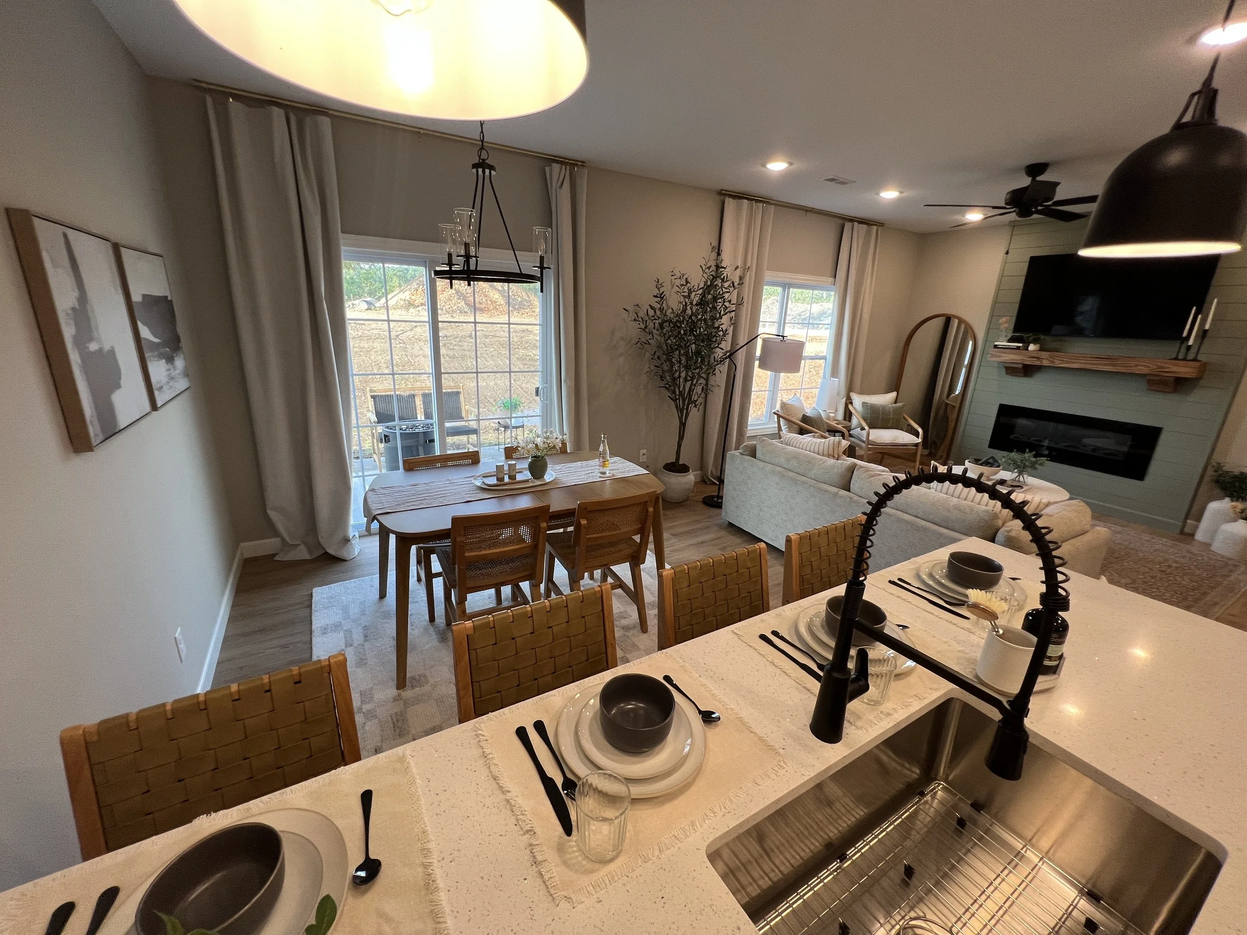 Open concept living and dining area with a kitchen island in the foreground. The island has place settings for four with black utensils, black bowls, and light-colored plates. A black kitchen faucet is visible over the stainless steel sink. The dinin