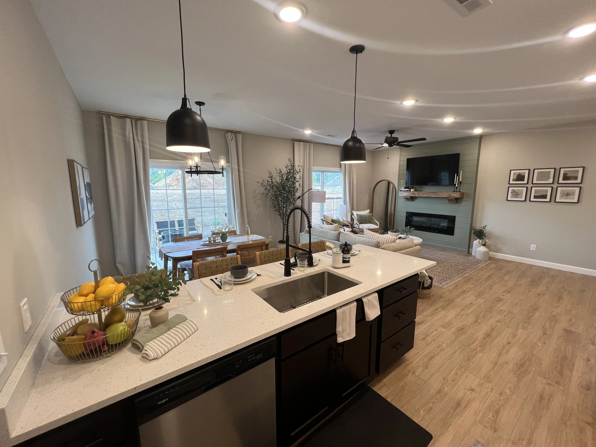 Open-concept living area with kitchen, dining, and sitting area. Kitchen with white countertop, black cabinets, fruit bowl, and black pendant lights. Dining area with wooden table and chairs. Living room with sofa, armchairs, fireplace, TV, and artwo