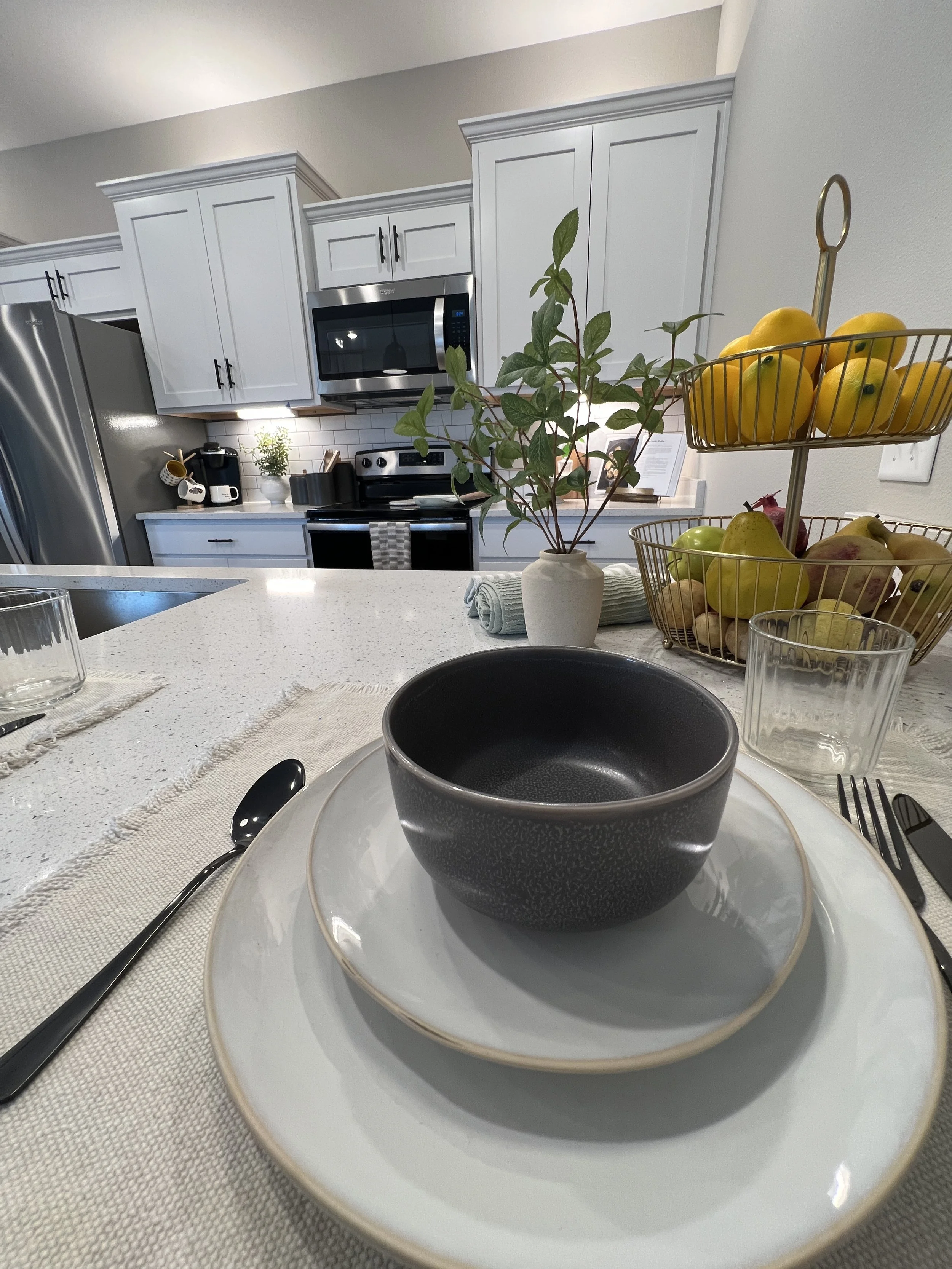 Decorative table setting in a modern kitchen with a ceramic bowl on a plate, glassware, cutlery, a potted plant, and fruit basket with lemons, apples, and pears.