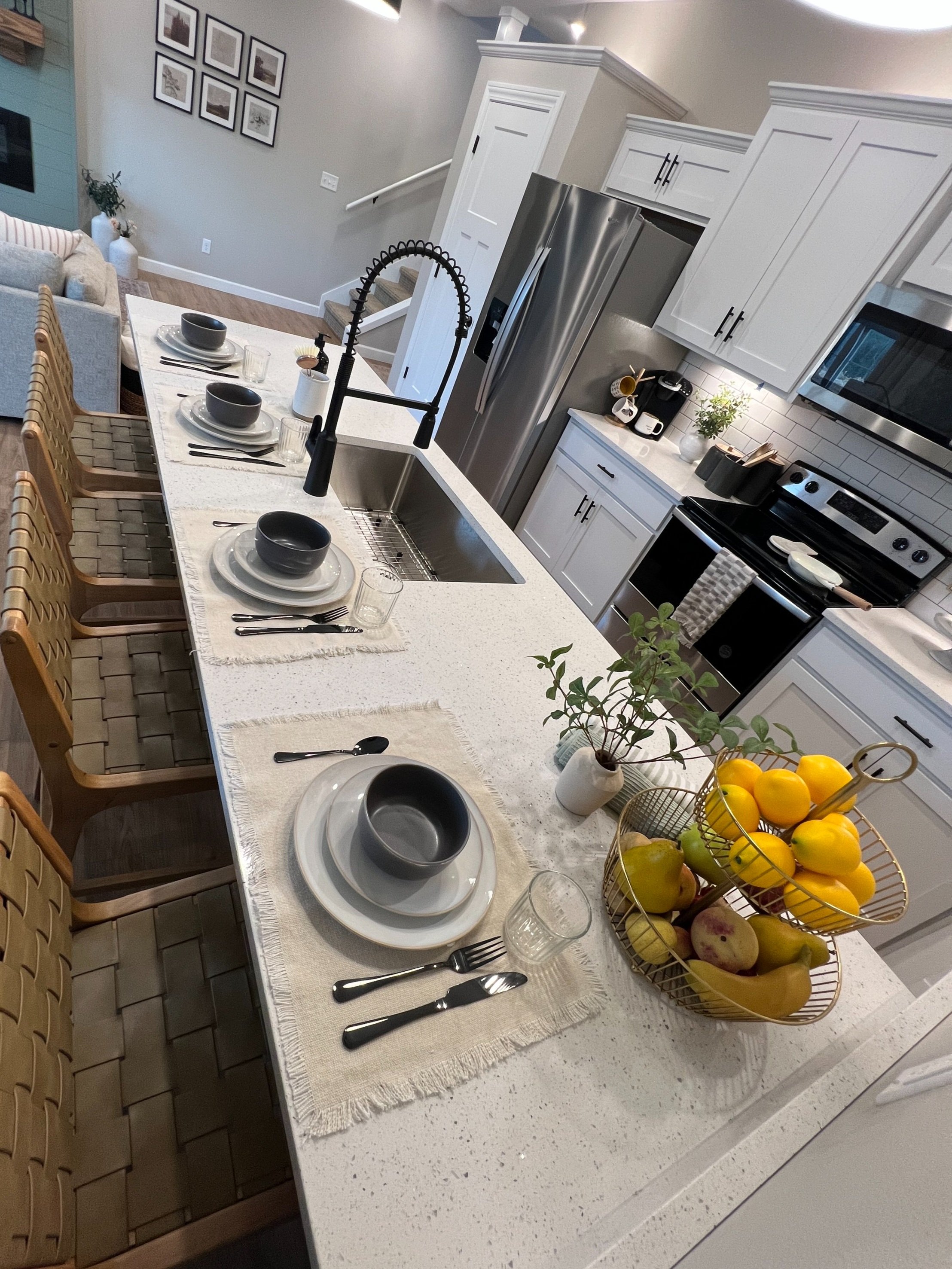 A modern kitchen with a white island counter set for four, with dark bowls, gray plates, and silverware. The counter has a built-in sink with a tall, black, flexible faucet. There are chairs with woven seats around the island. The kitchen has white c