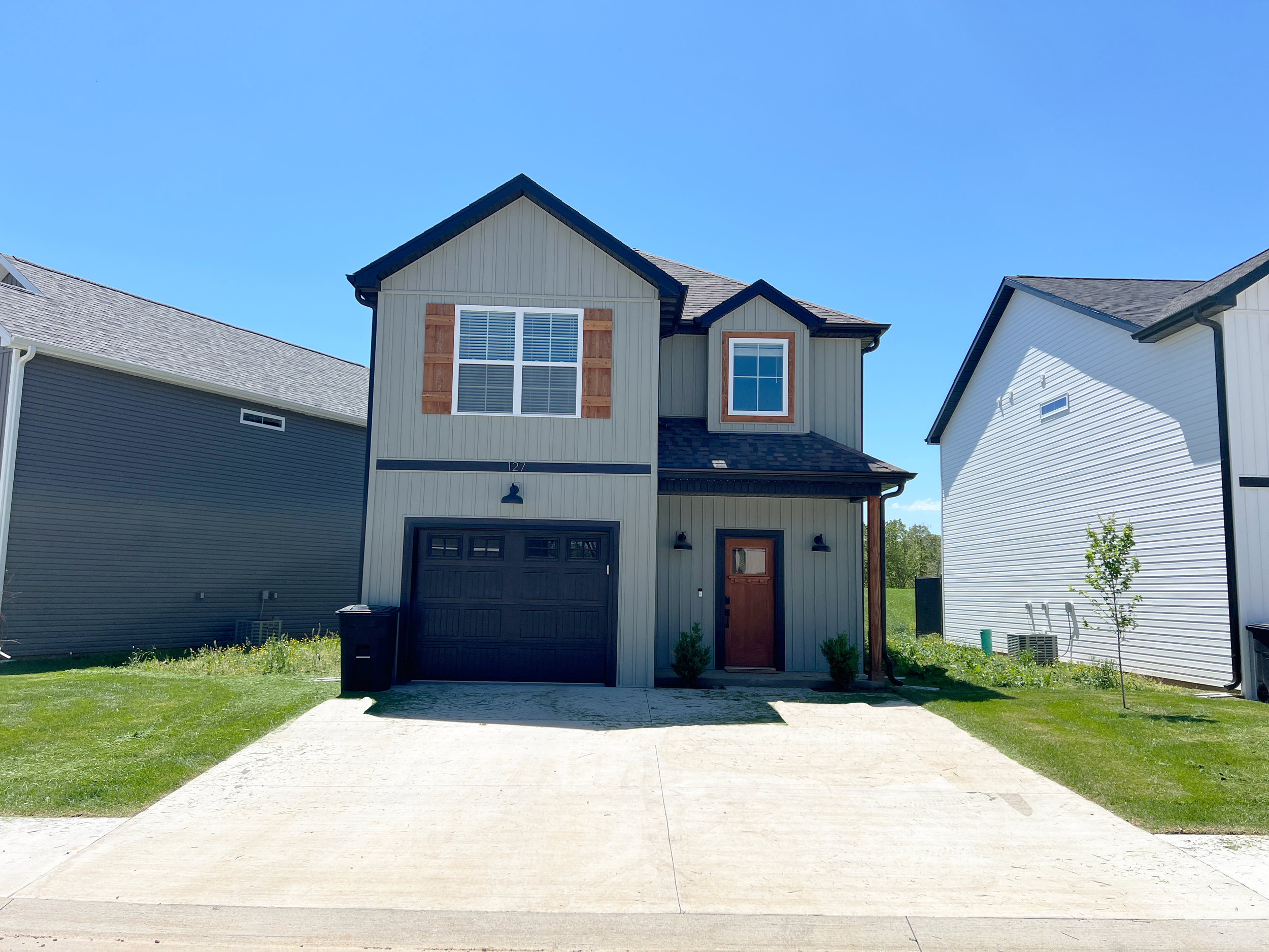 Front view of a modern two-story house with a garage, a wooden front door, and siding painted in beige and black accents, flanked by neighboring houses and a clear blue sky.