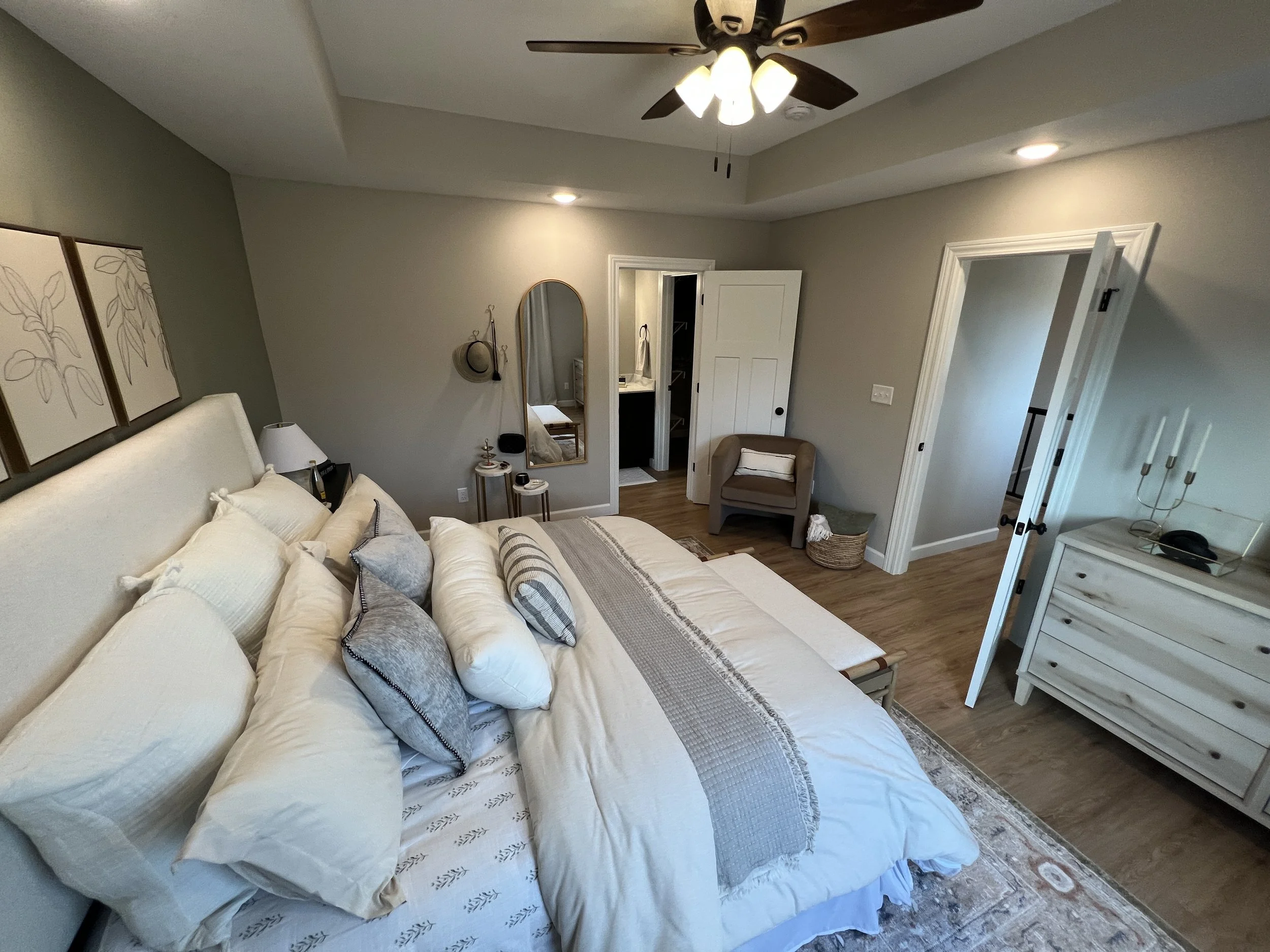 A cozy bedroom with a large bed covered in cream and gray bedding and multiple pillows, a ceiling fan with lights, a small chair in the corner, a dresser with a candle holder and decorative items, a mirror on the wall, and an open door leading to a b