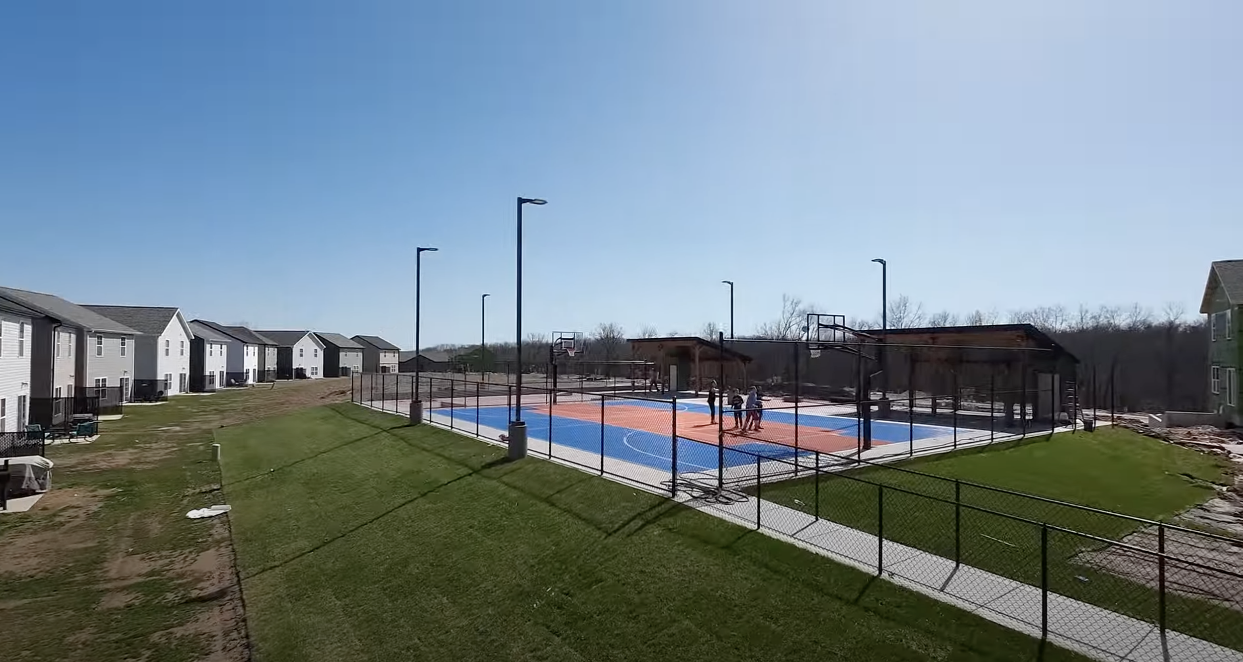 Newly constructed outdoor basketball court with blue and orange surface, enclosed by black chain-link fencing, situated near a row of residential buildings under a clear blue sky.