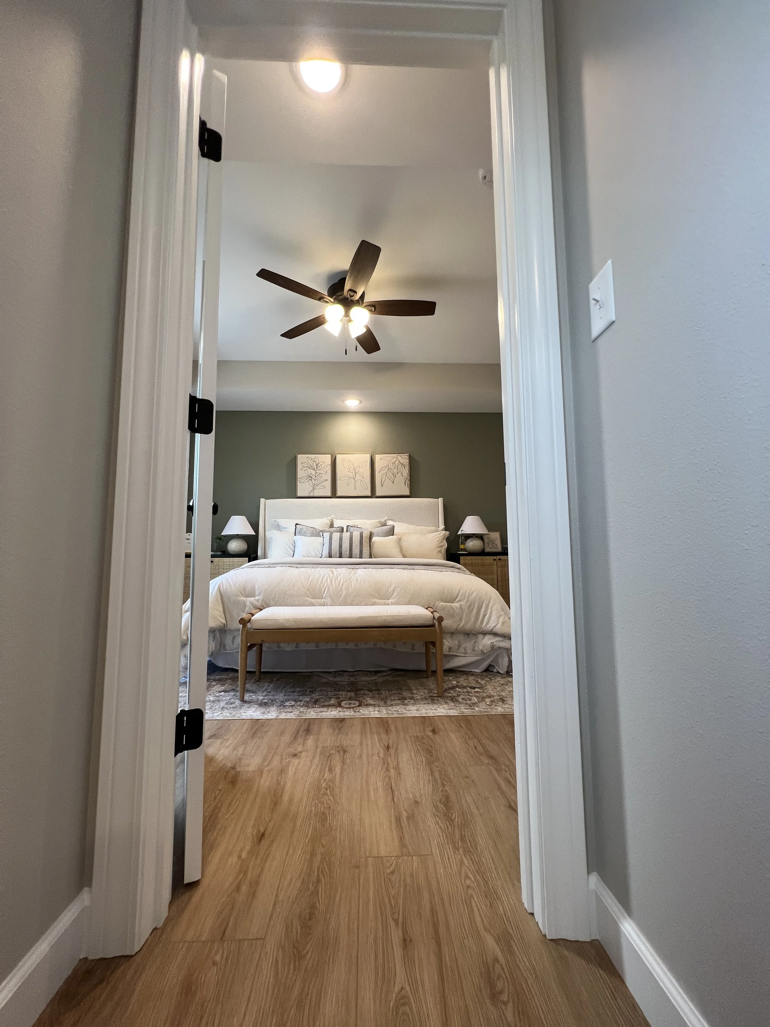 View through a doorway into a bedroom with a bed, nightstands, lamps, and wall art, featuring a ceiling fan and wood flooring.
