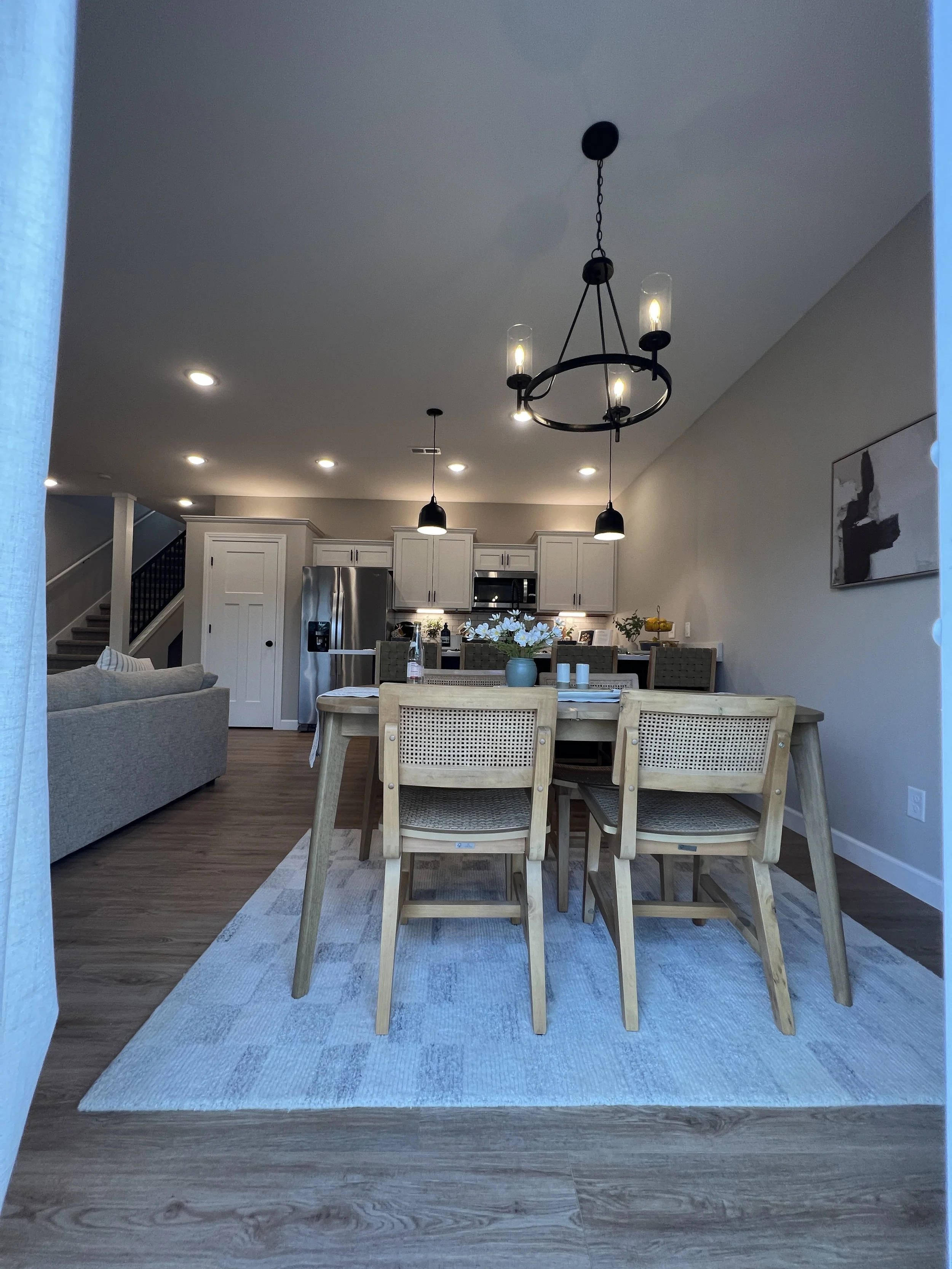 View of a dining area with a wooden table, matching chairs, an area rug, a chandelier, and a kitchen in the background with white cabinets and a stainless steel refrigerator.