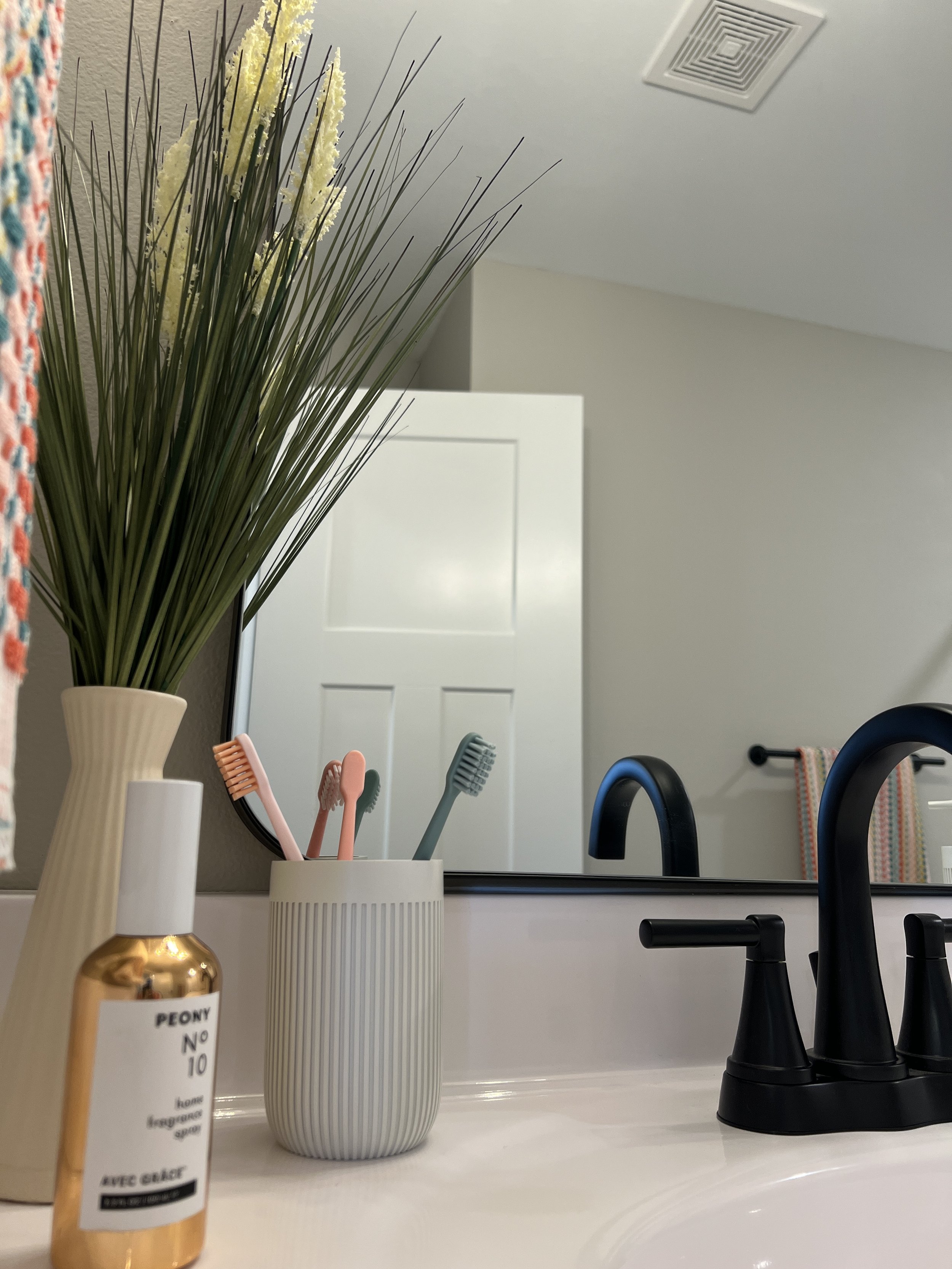 Bathroom sink area with a large mirror, black faucet, and soap dispenser. Decorative items include a tall beige vase with artificial grass and yellow flowers, a white ribbed cup holding three toothbrushes in pink, peach, and green. In the background,