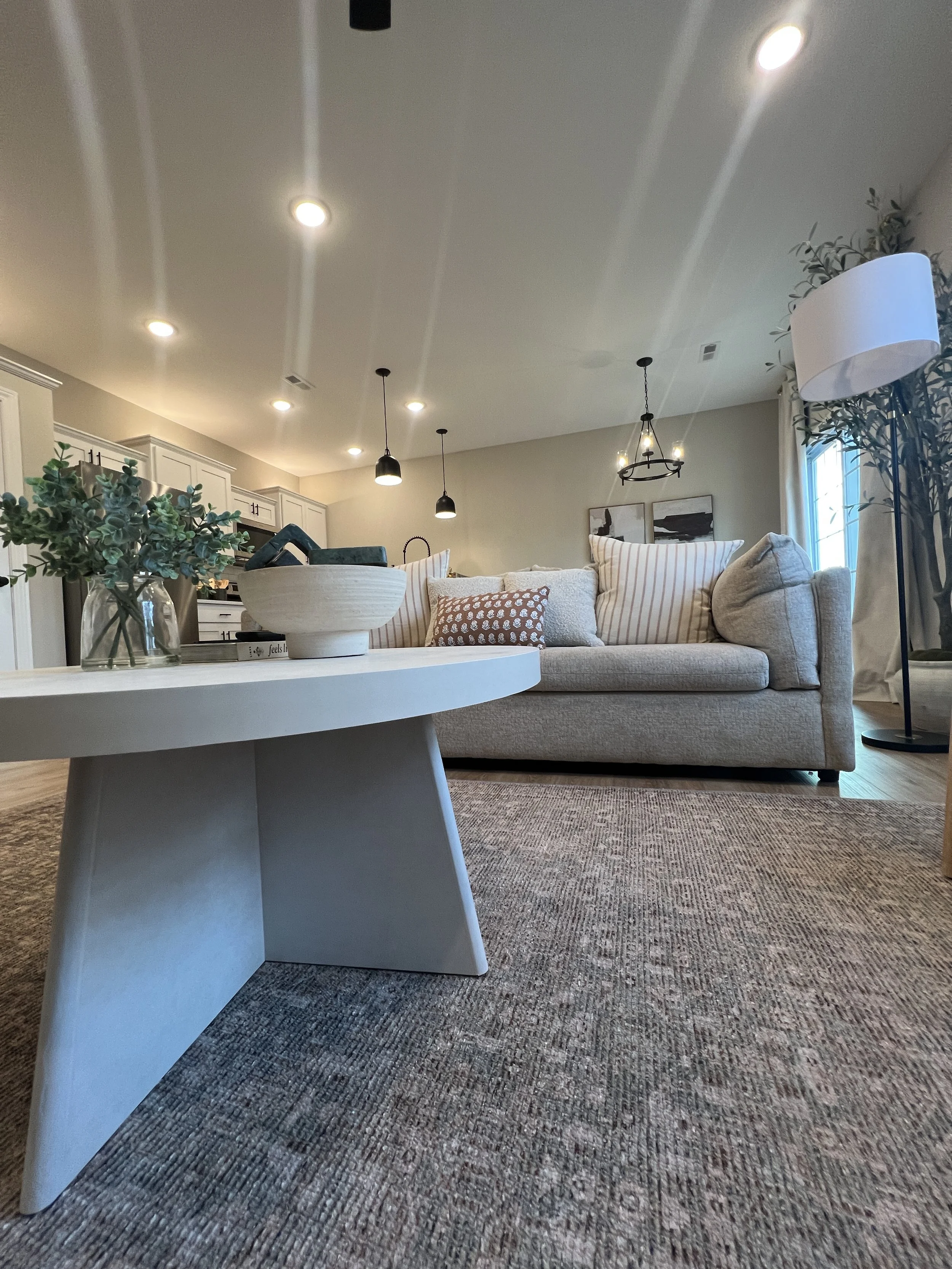Living room with beige sofa, white coffee table, and potted plants, illuminated by ceiling and pendant lights.