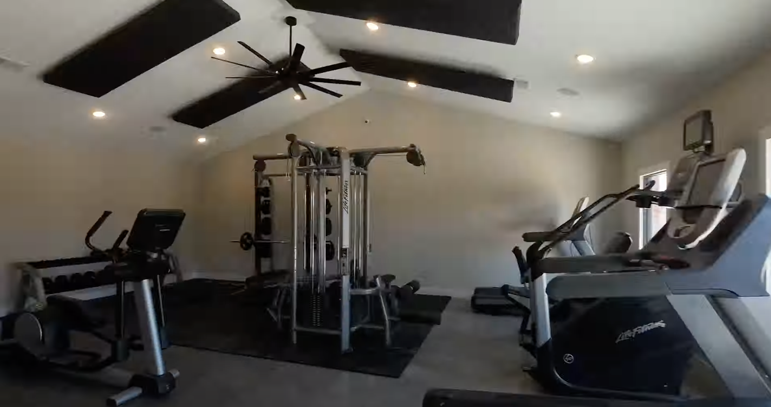Interior view of a home gym with exercise equipment including a treadmill, elliptical, and weight machine, with black acoustic panels on the ceiling and recessed lighting.