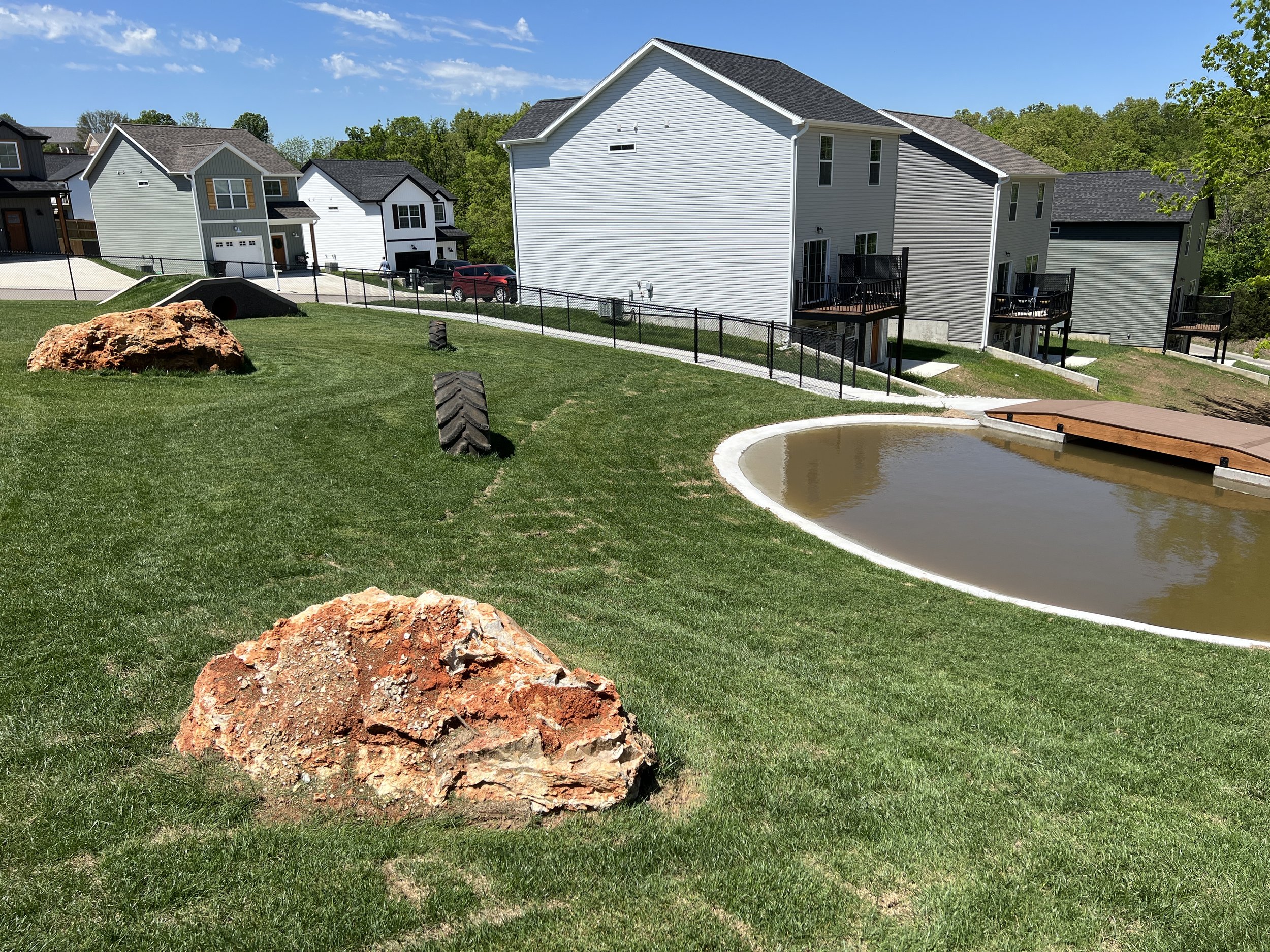 Residential backyard with grassy lawn, large rocks, a small pond, and multiple houses with decks in the background.