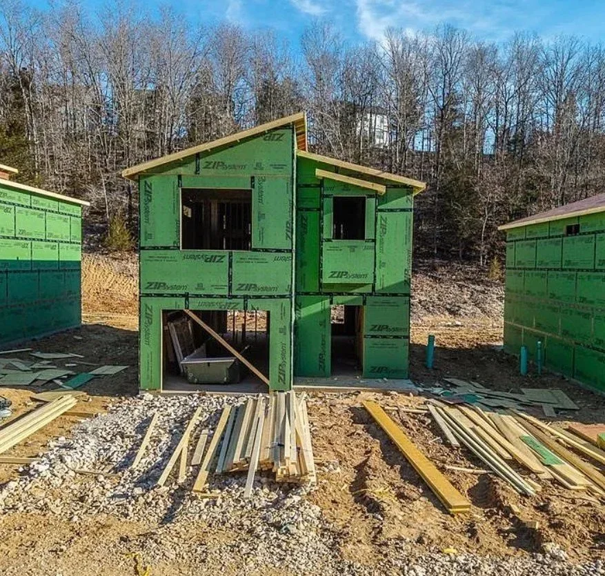 Under construction two-story house with green sheathing panels on the exterior, setting on a gravel foundation, with piles of lumber and construction materials on the ground