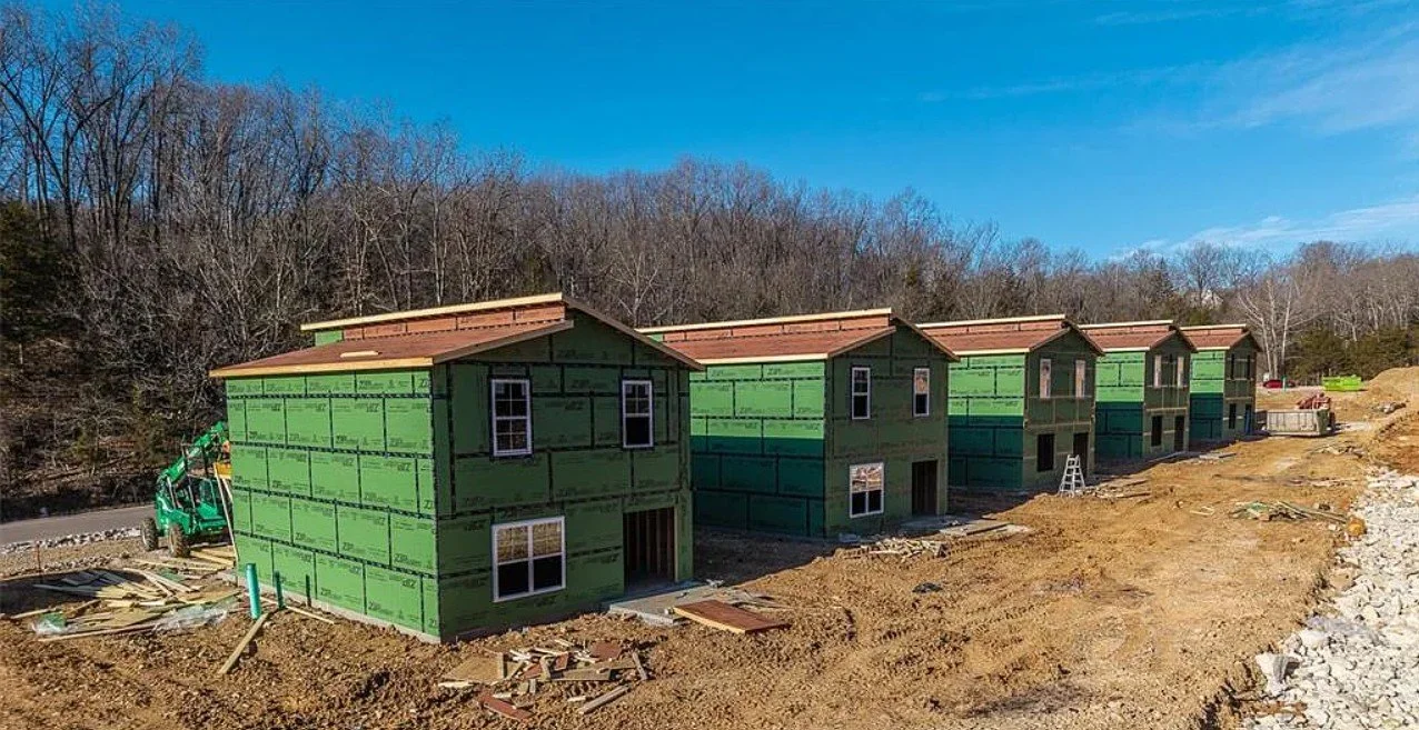 Several green residential buildings under construction with a wooded hillside in the background and a dirt area in the foreground.