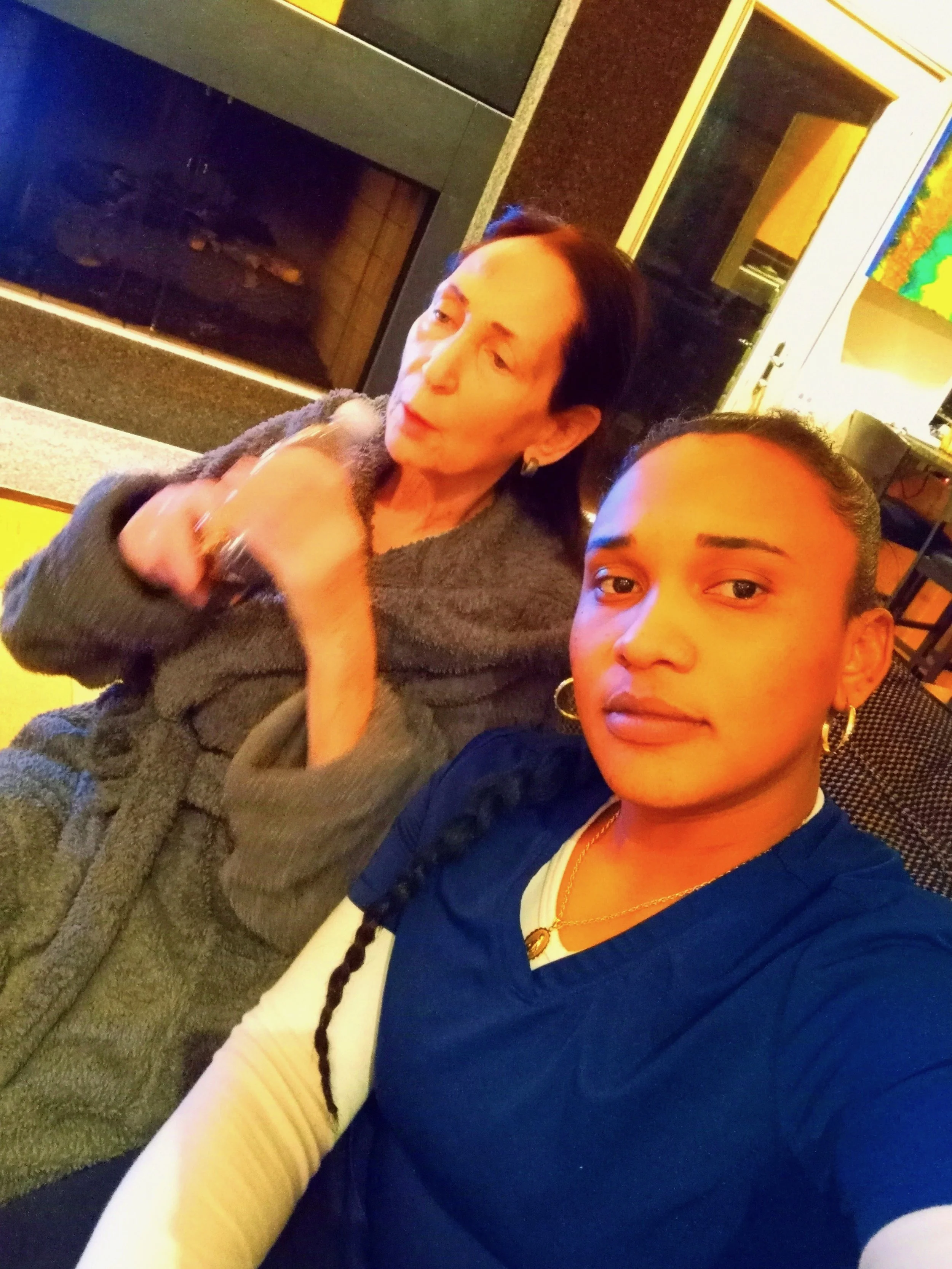Two women sitting in a living room with a fireplace, taking a selfie. One woman has dark hair styled in a braid, wearing a blue top with white sleeves, and gold jewelry. The other woman has short dark hair, wearing a gray sweater, and holding her hand near her face.