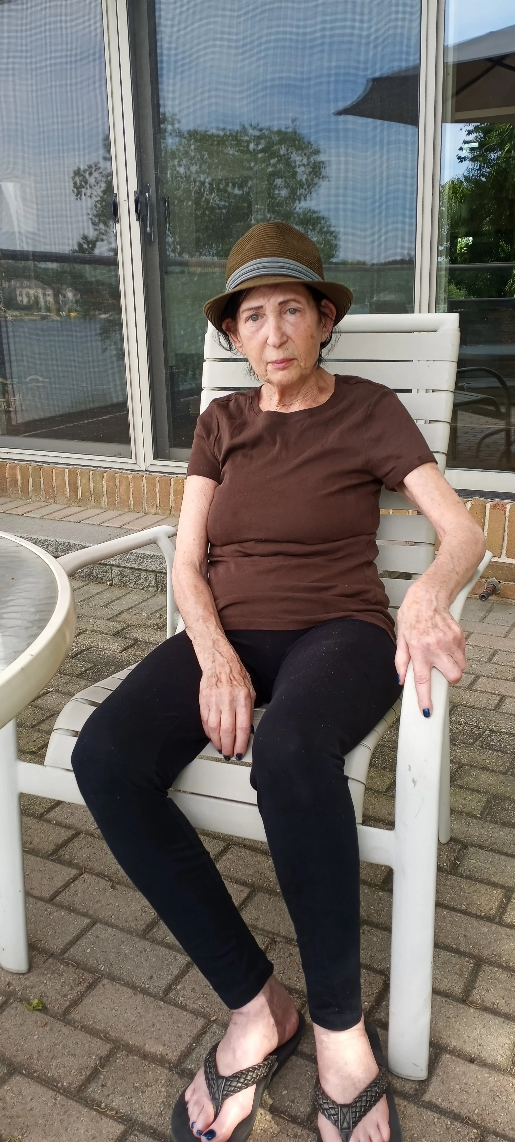 An elderly woman sitting on a white chair outdoors, wearing a brown shirt, black pants, sandals, and a brown hat with a ribbon, with a reflective glass window behind her.