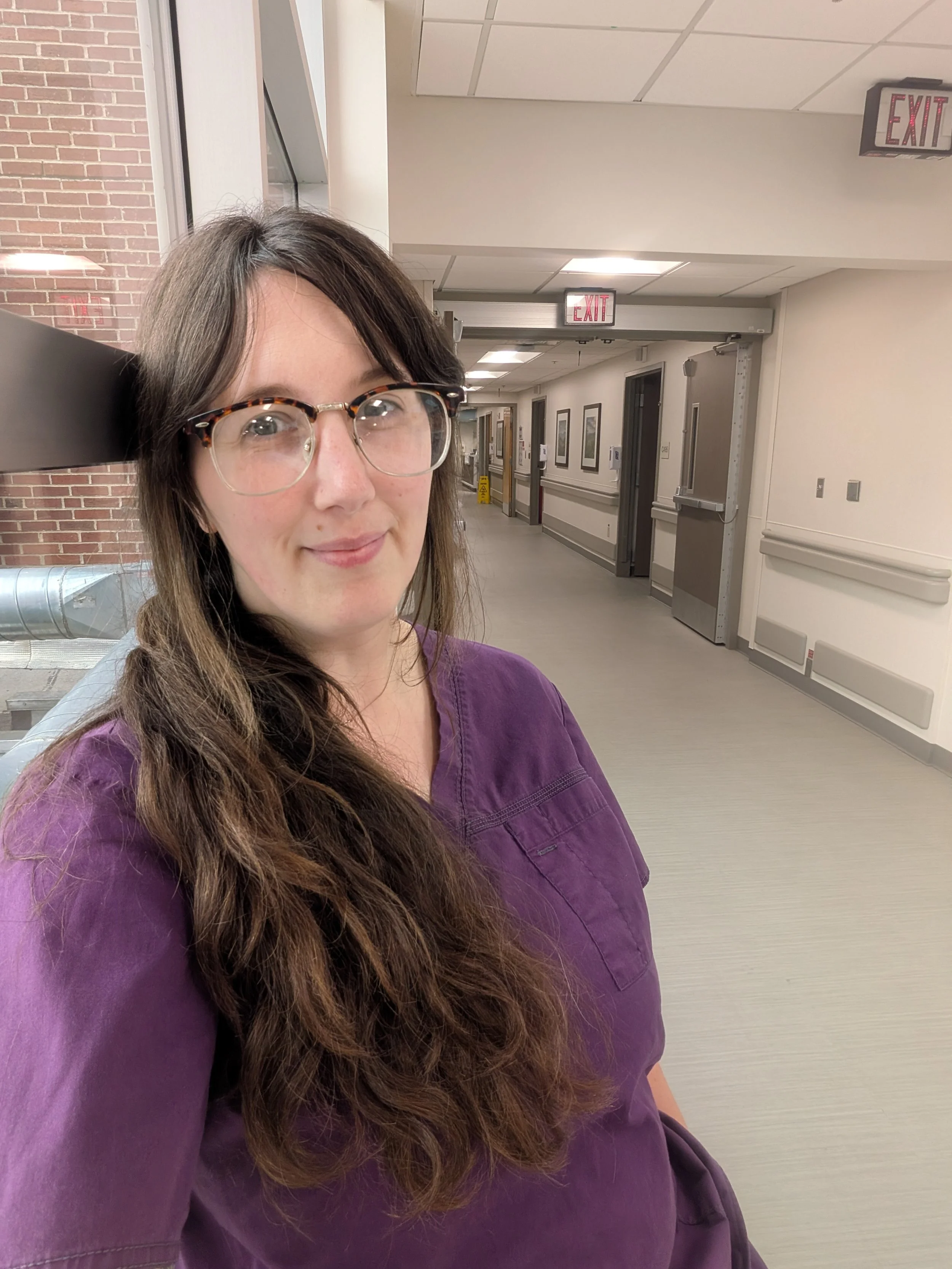 A woman with is standing in a hallway at Mission Hospital in her purple doula scrubs