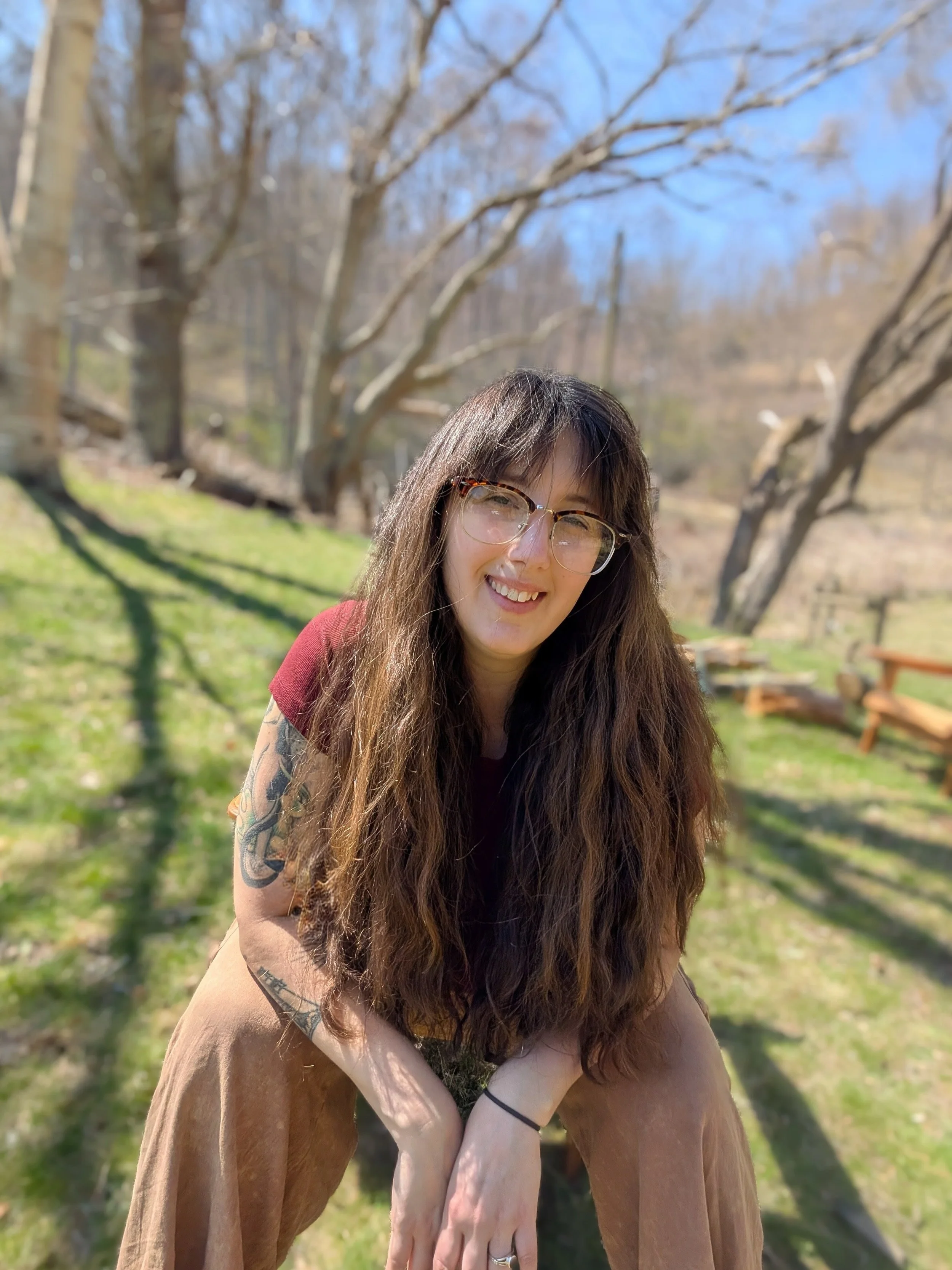 A woman with long wavy brown hair, wearing glasses, a maroon shirt, and tan pants, smiling outdoors on a sunny day with a background of leafless trees and green grass.