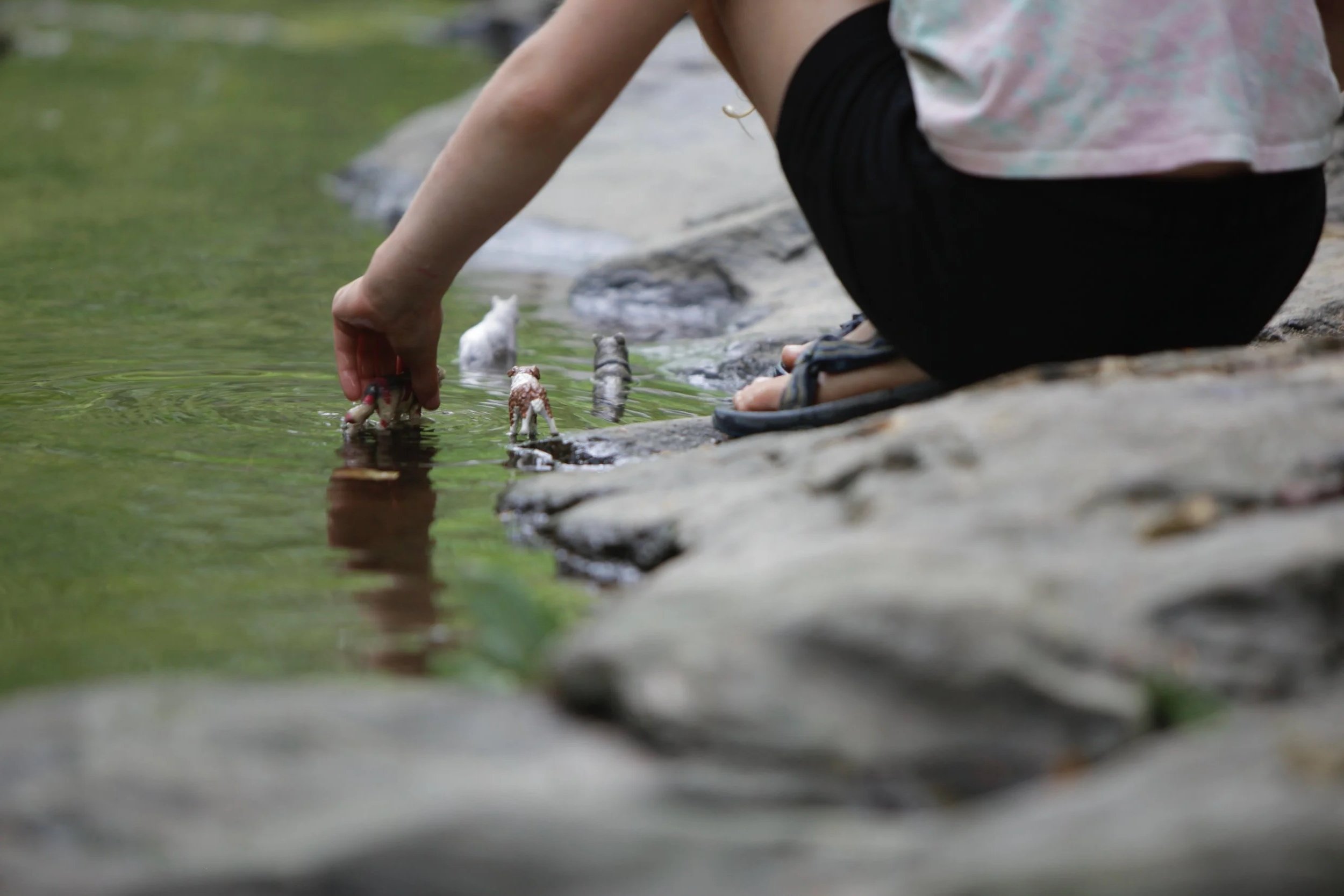 Child crouching by a creek, reaching into the water, with small toy animals and rocks visible on the creek bank.