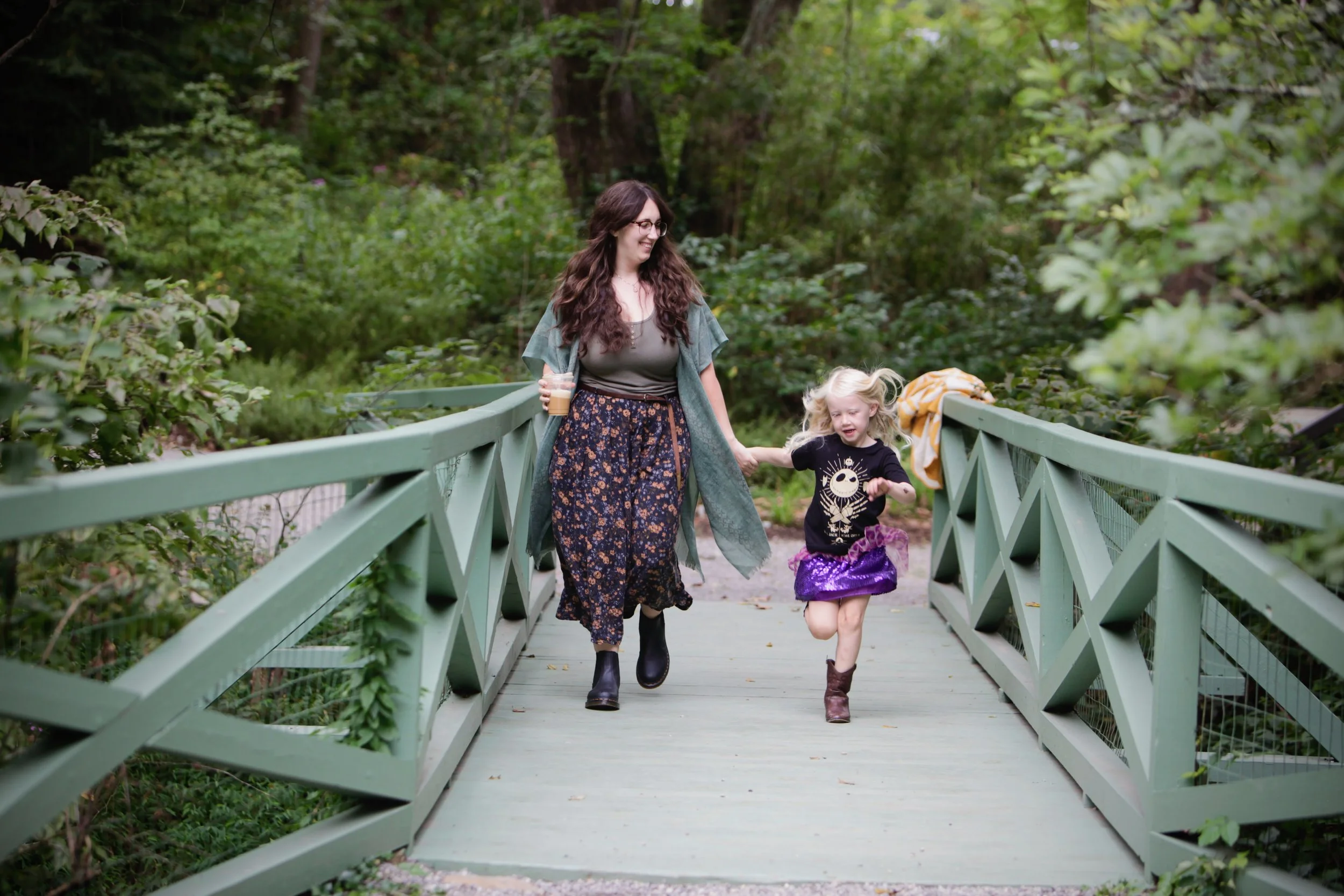 A woman and a young girl walking on a green wooden bridge in a lush forest, holding hands, with the girl skipping and the woman smiling.