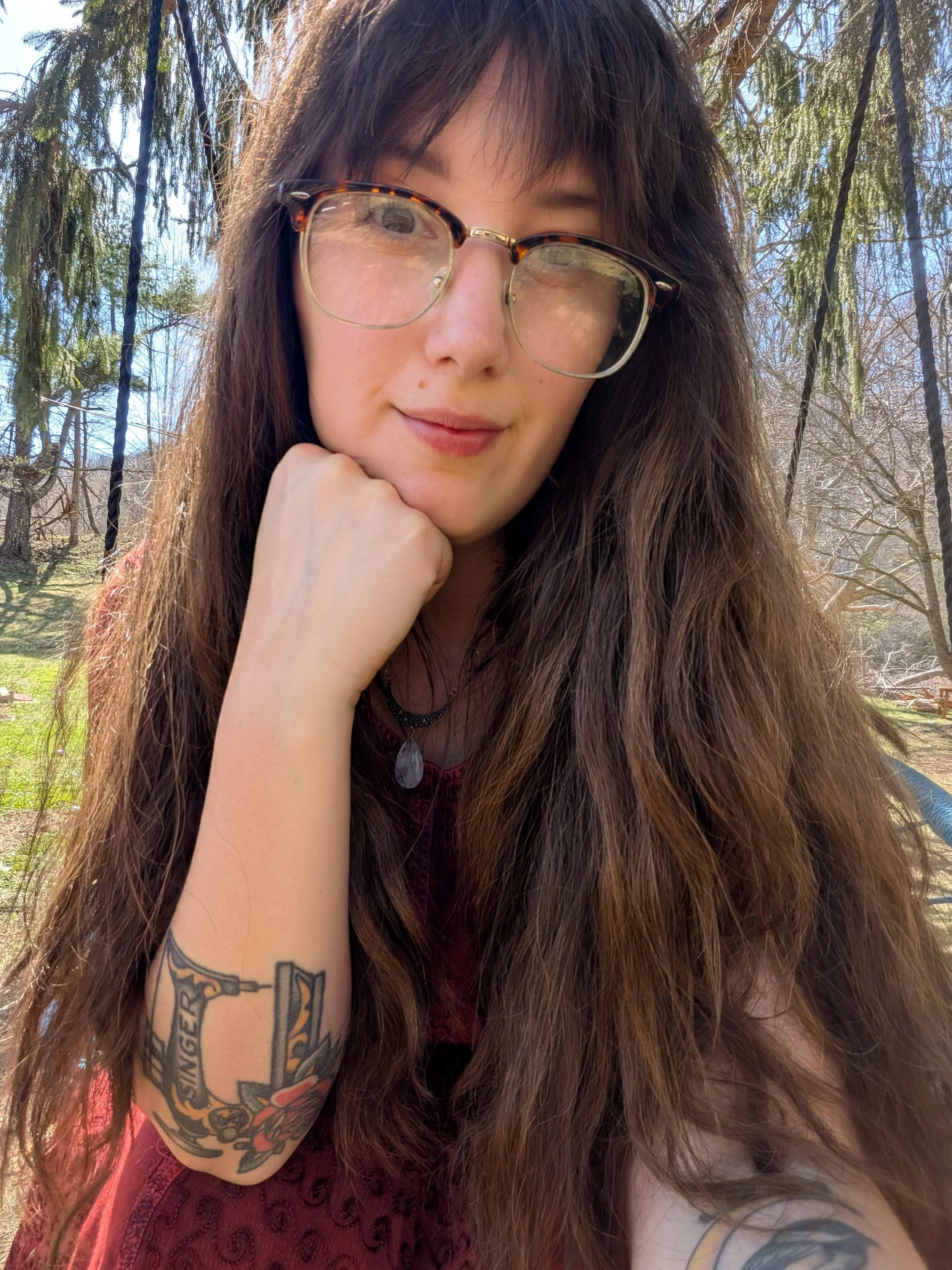 A woman with long brown hair and glasses, sitting outdoors with trees and a blue sky in the background, resting her chin on her hand and smiling.