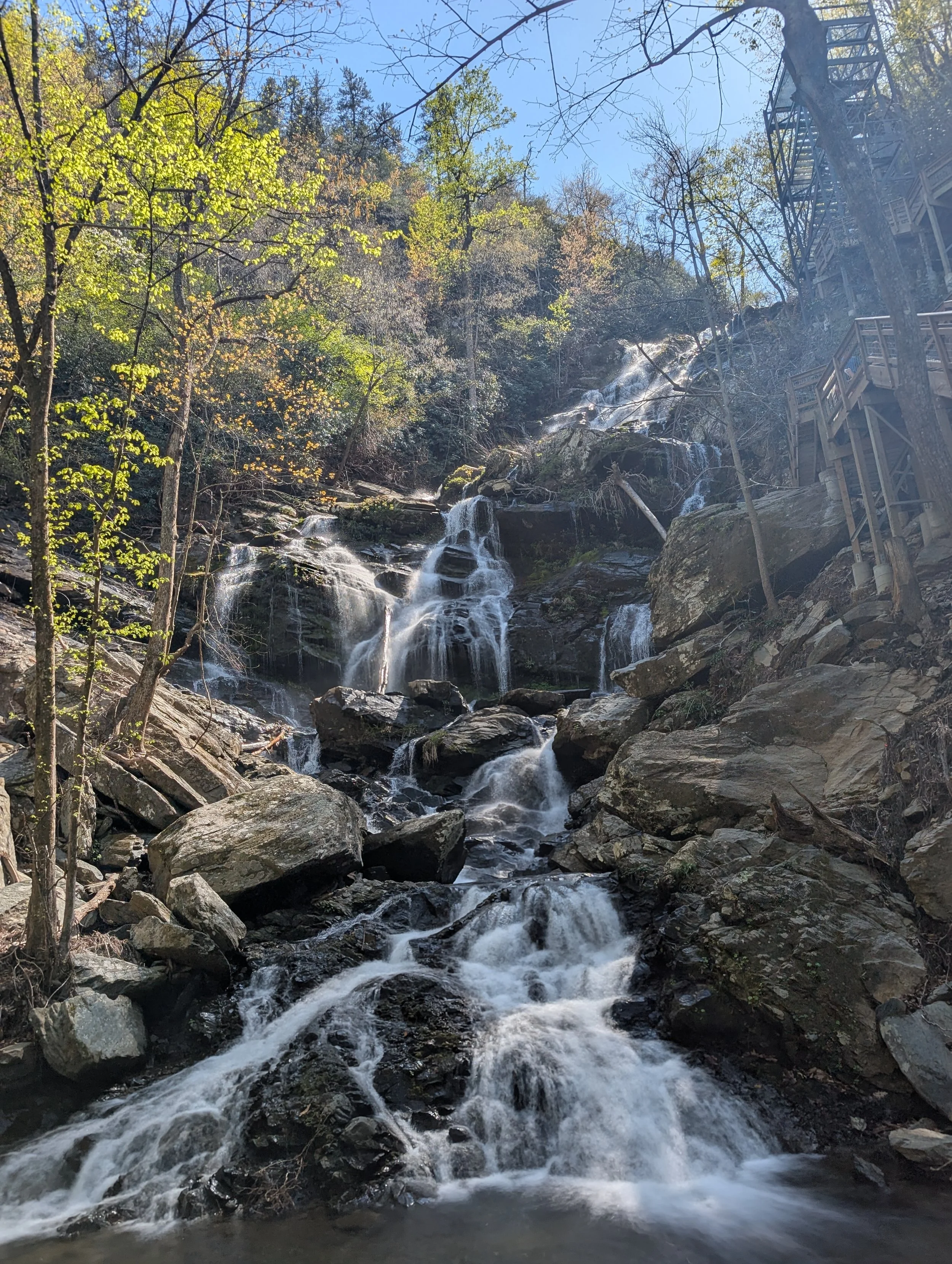 A scenic outdoor waterfall cascading down rocks surrounded by trees with some leaves beginning to change color and a wooden staircase or platform on the right side.
