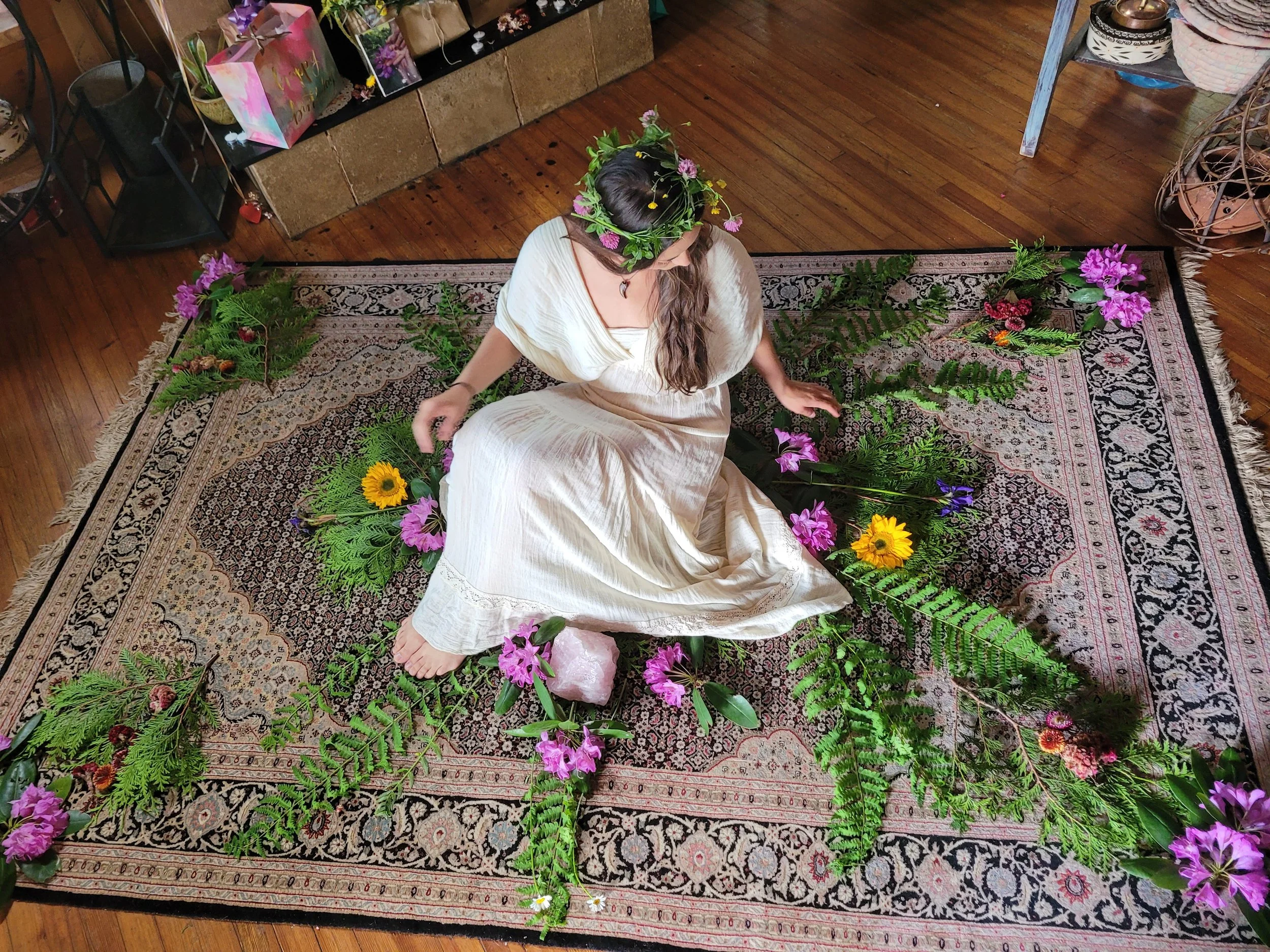 A woman wearing a white dress and a flower crown sits on a decorative rug surrounded by various colorful flowers and green fern leaves, with a wooden floor underneath.