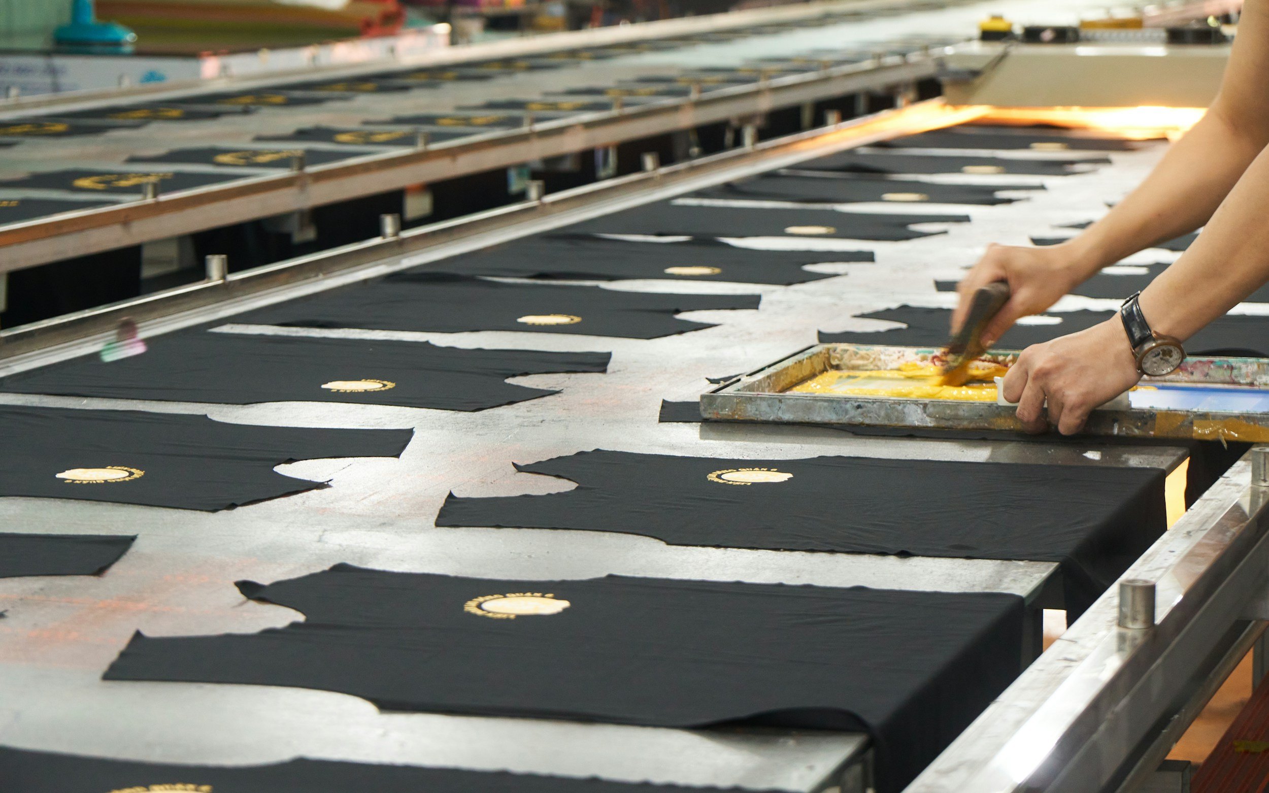 Hands working on screen printing black shirts with a gold logo on a printing press.