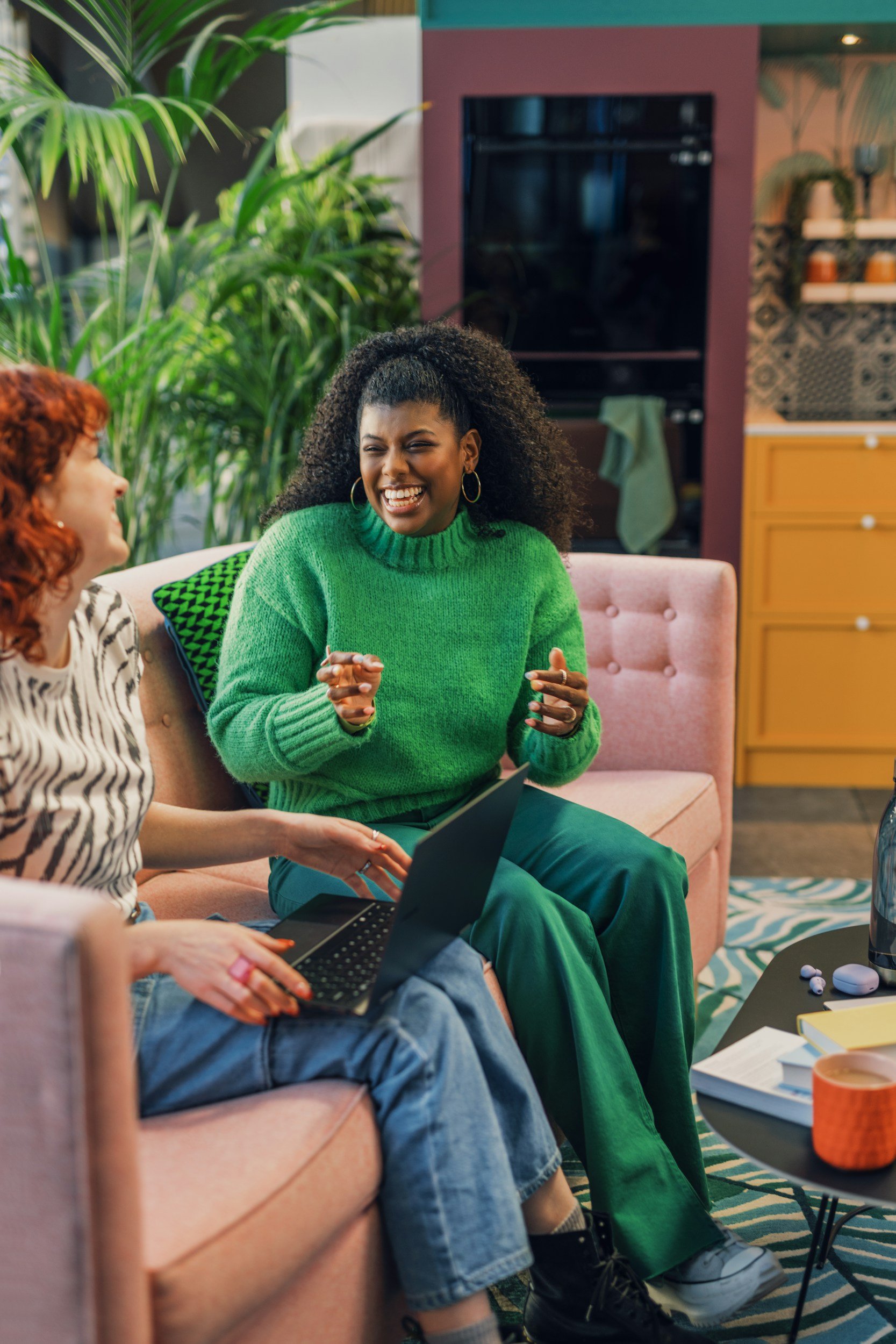 Two women are sitting on a pink sofa, laughing and talking in a colorful, cozy living room. One woman is wearing a green sweater, and the other has red hair and is using a laptop.