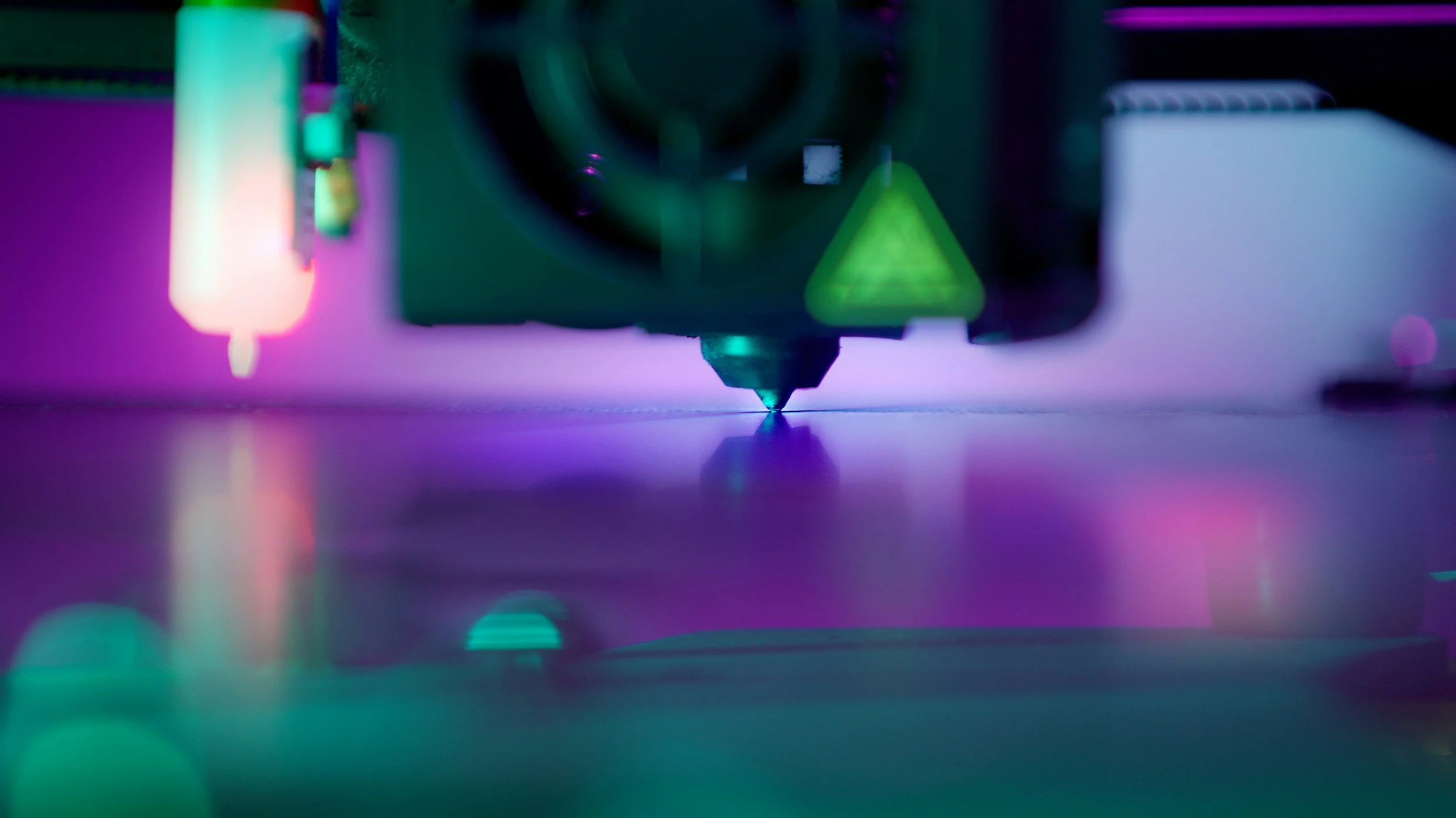 Close-up of a 3D printer nozzle extruding purple filament, with colorful LED lighting creating a vibrant glow around the process