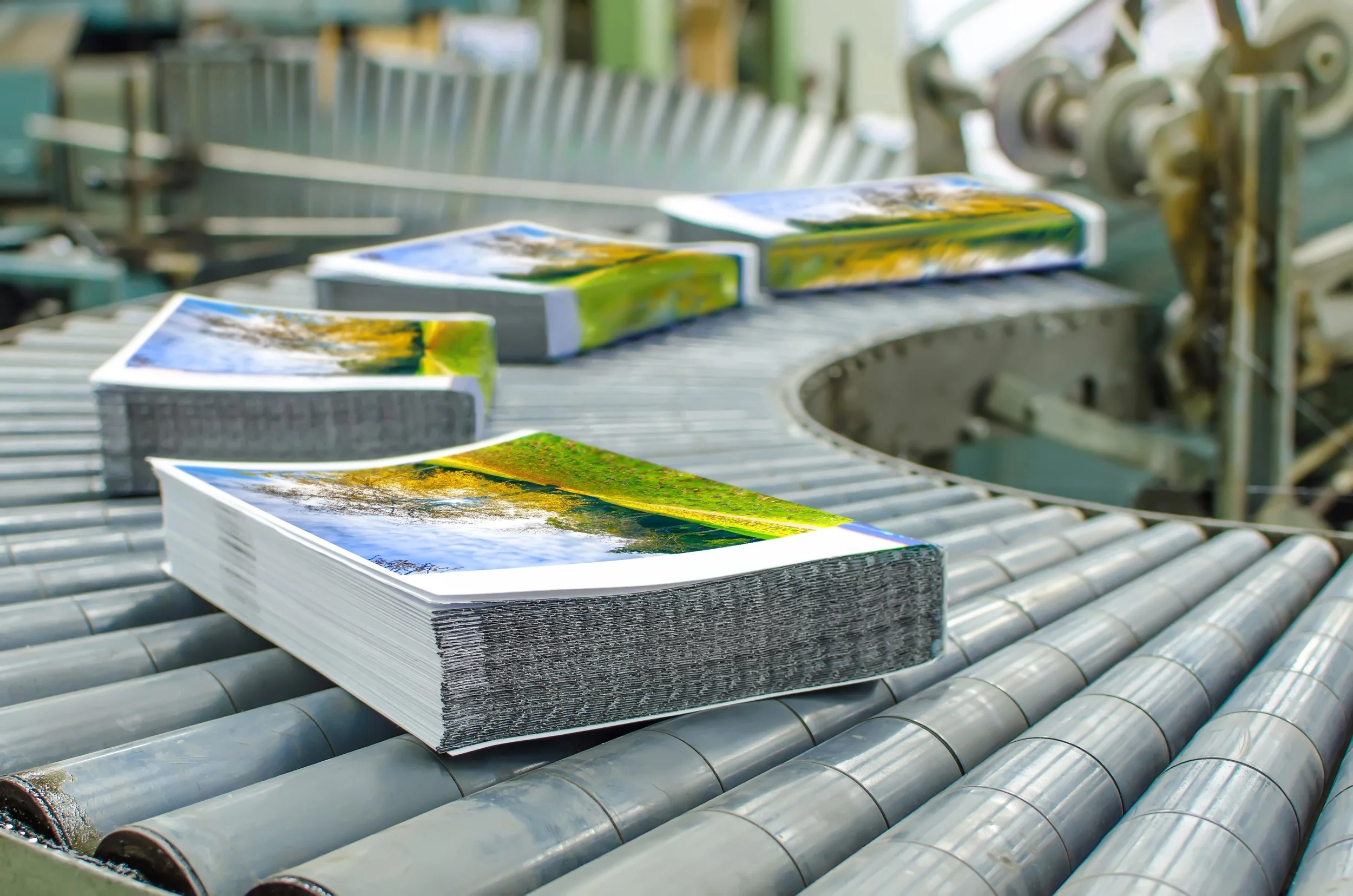 Photographs of outdoor scenes on stacks of printed brochures or magazines on a conveyor belt in a production or printing facility.