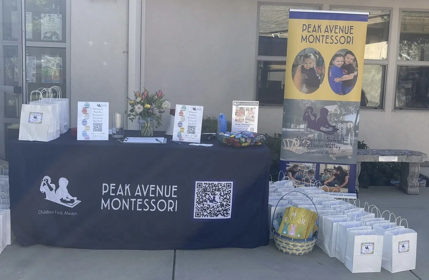 Outdoor event booth for Peak Avenue Montessori with a black tablecloth, informational signs, a flower arrangement, and gift bags. A banner displays three children, the school's name, and logo. The table has a QR code and a basket of colorful items, w