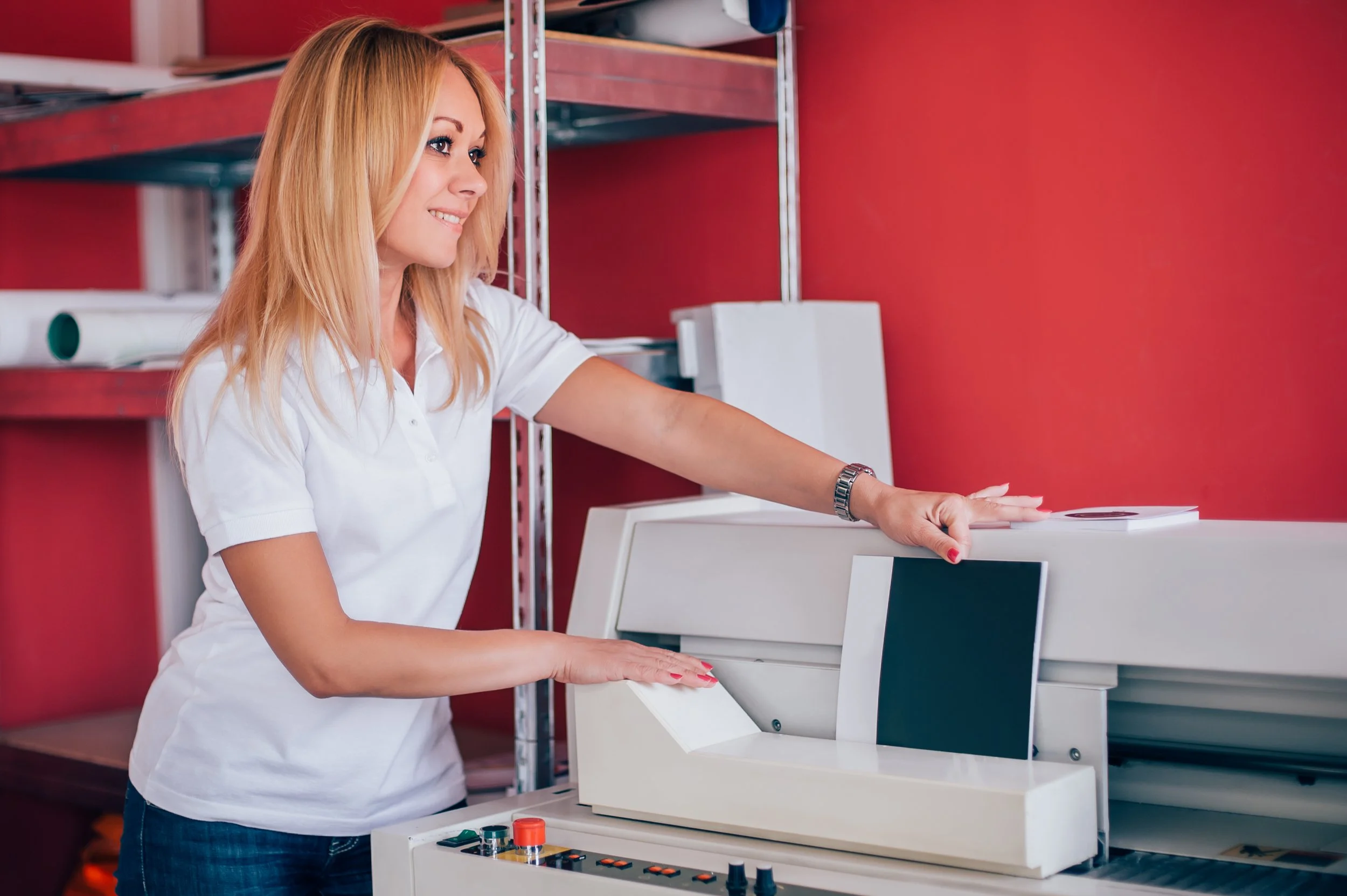 A woman with blonde hair and a white shirt operating a large, vintage electronic device with a screen and controls in a room with red walls and shelving units.