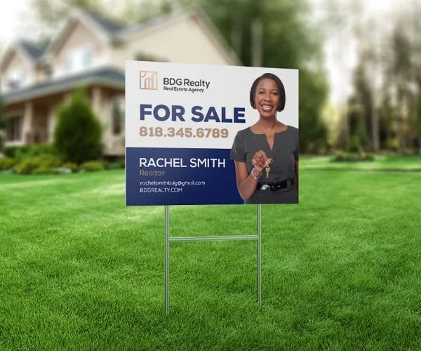 A real estate sign for sale on a lawn in front of a house, featuring a smiling woman holding a key, with contact information for Rachel Smith, a realtor at BDG Realty.