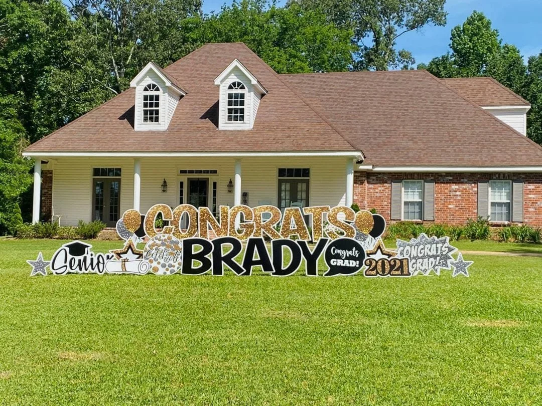 Graduation congratulatory decorations on a lawn in front of a house, with signs saying 'Congrats Brady,' 'Senior,' 'Grad,' and the year 2021, featuring stars, graduation cap, and diploma graphics.