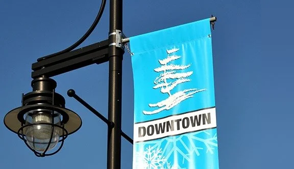 Downtown streetlamp with a bright blue banner featuring a white tree and snowflake design, under a clear blue sky.