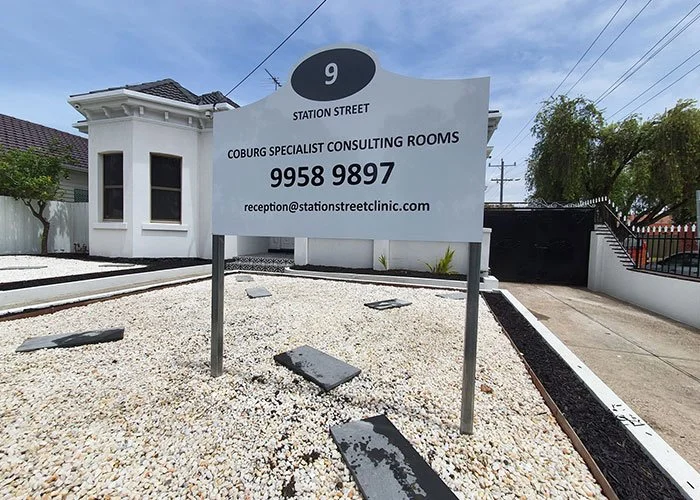 Sign for Station Street, Coburg Specialist Consulting Rooms, with contact number and email, in front of a white building and trees.