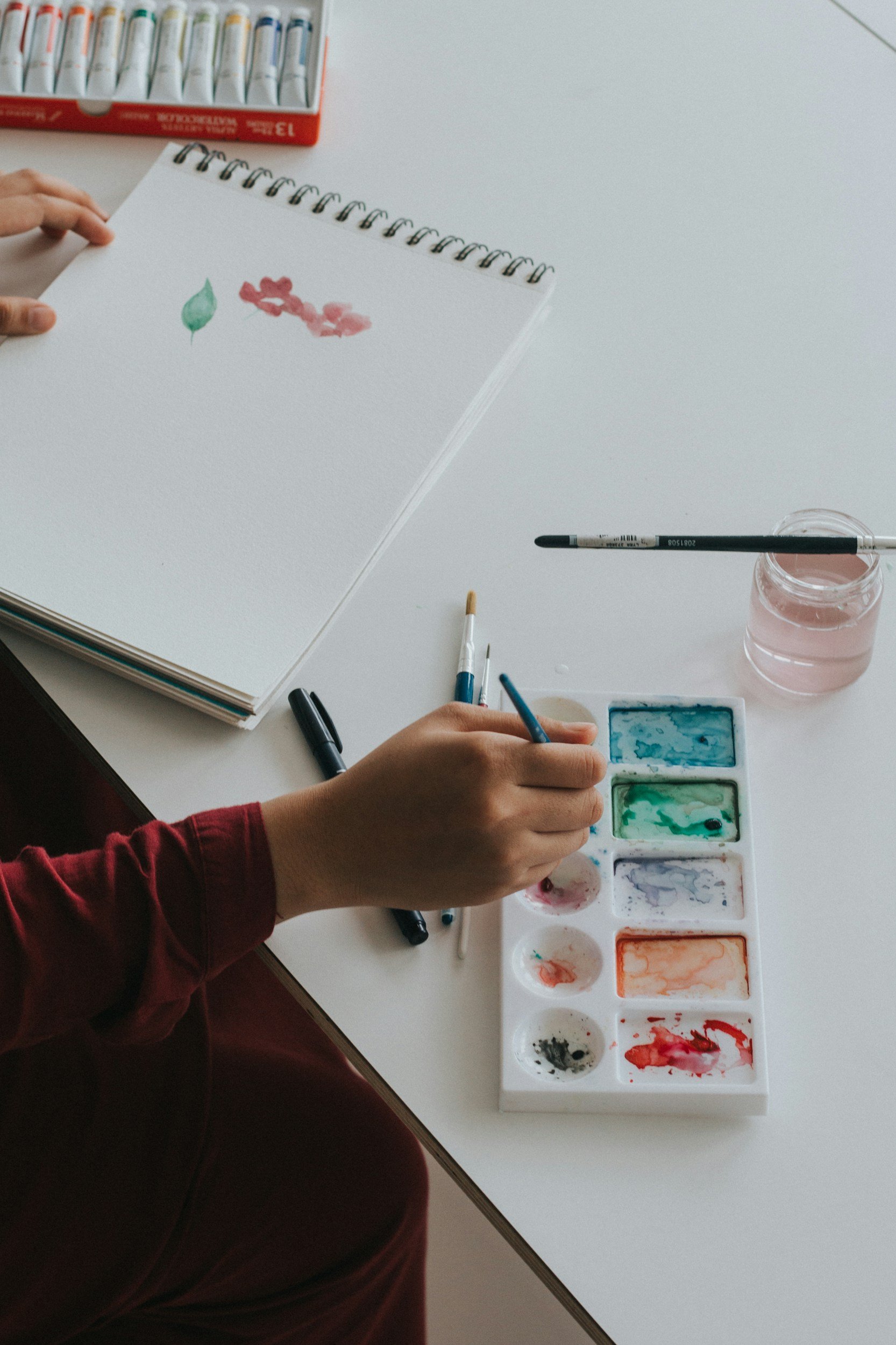 Person painting with watercolors on a white notebook page. Art supplies including watercolor palette, brushes, and pens are on the table.