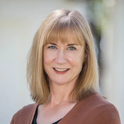Portrait of a woman with blonde hair, blue eyes, and a friendly smile, wearing a brown and black top, outdoors with blurred background.