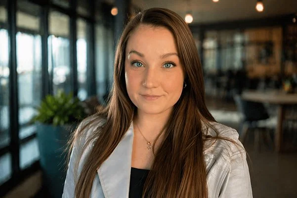 A young woman with long brown hair and blue eyes looks into the camera. She wears a light-colored jacket and a delicate necklace, with a calm expression. Blurred background.