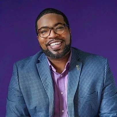 Smiling man in glasses and a purple shirt with a checked blazer against a purple background, conveying a friendly and approachable tone.