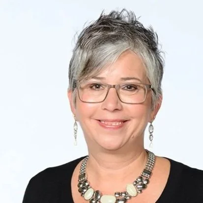 A woman with short gray hair smiles warmly at the camera. She wears glasses, a black top, dangling earrings, and a chunky beaded necklace, exuding confidence.
