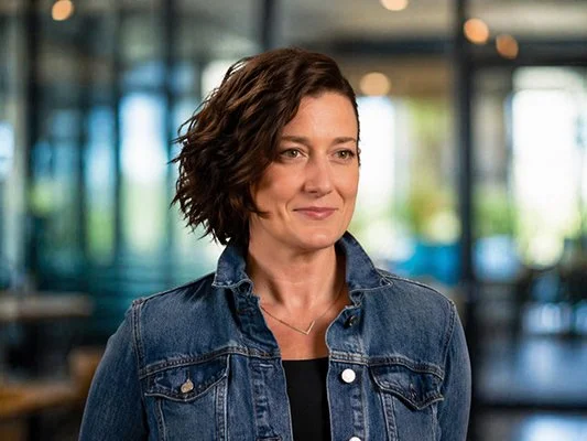 Smiling woman with short, wavy brown hair in a denim jacket, standing in a softly blurred indoor setting, exuding a confident and warm demeanor.