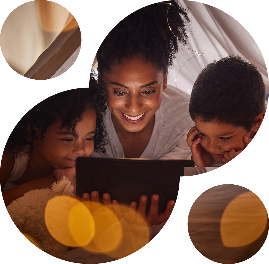 A woman and two children, smiling, watch a tablet together under a cozy blanket fort, creating a warm, intimate atmosphere with soft lighting.