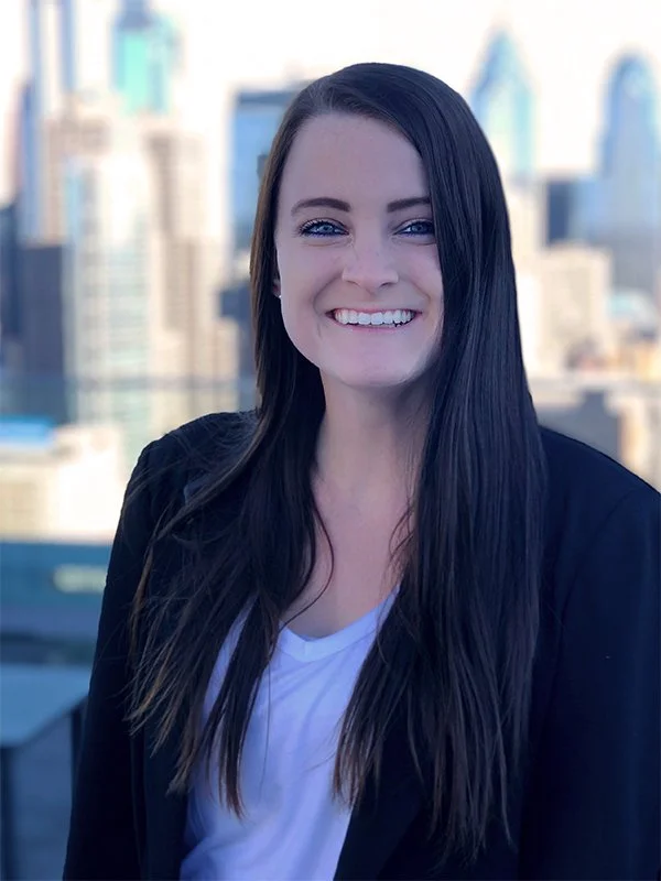 Smiling woman with long brown hair stands outdoors against a blurred urban skyline, wearing a black blazer and white shirt, conveying a cheerful mood.
