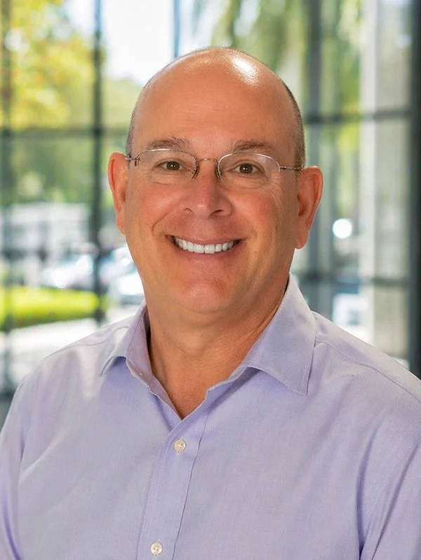 A smiling man in glasses and a light purple shirt is indoors with a blurred glass window and greenery in the background, conveying a professional tone.